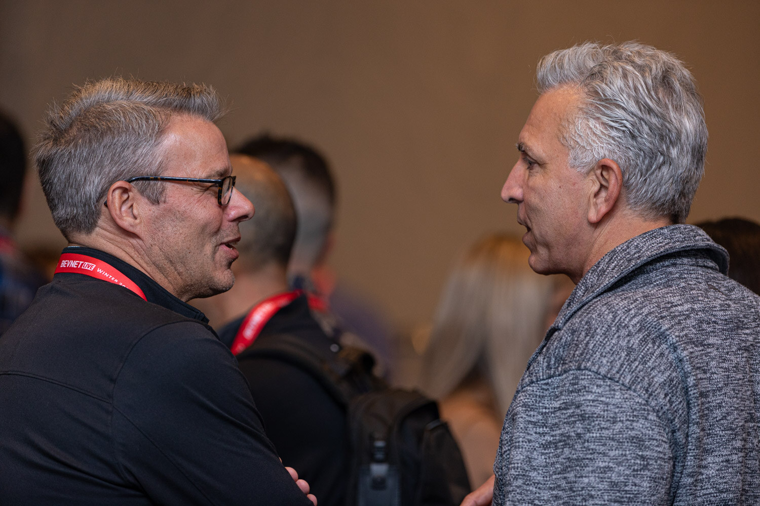 Two men with lanyards stand close and talk in a dimly lit hallway.