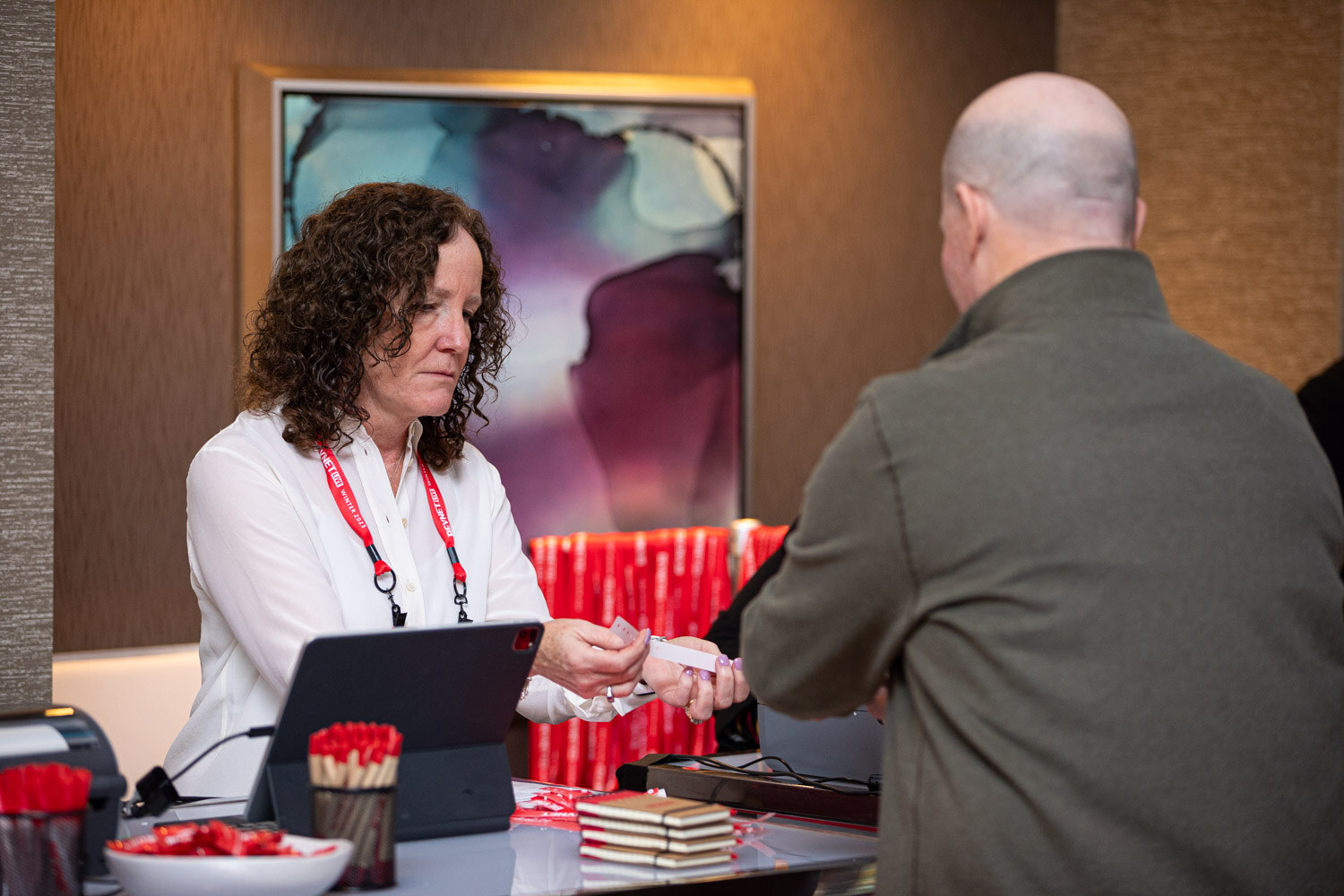 A woman with curly hair in a white shirt hands an item to a man across a registration table with red pens.