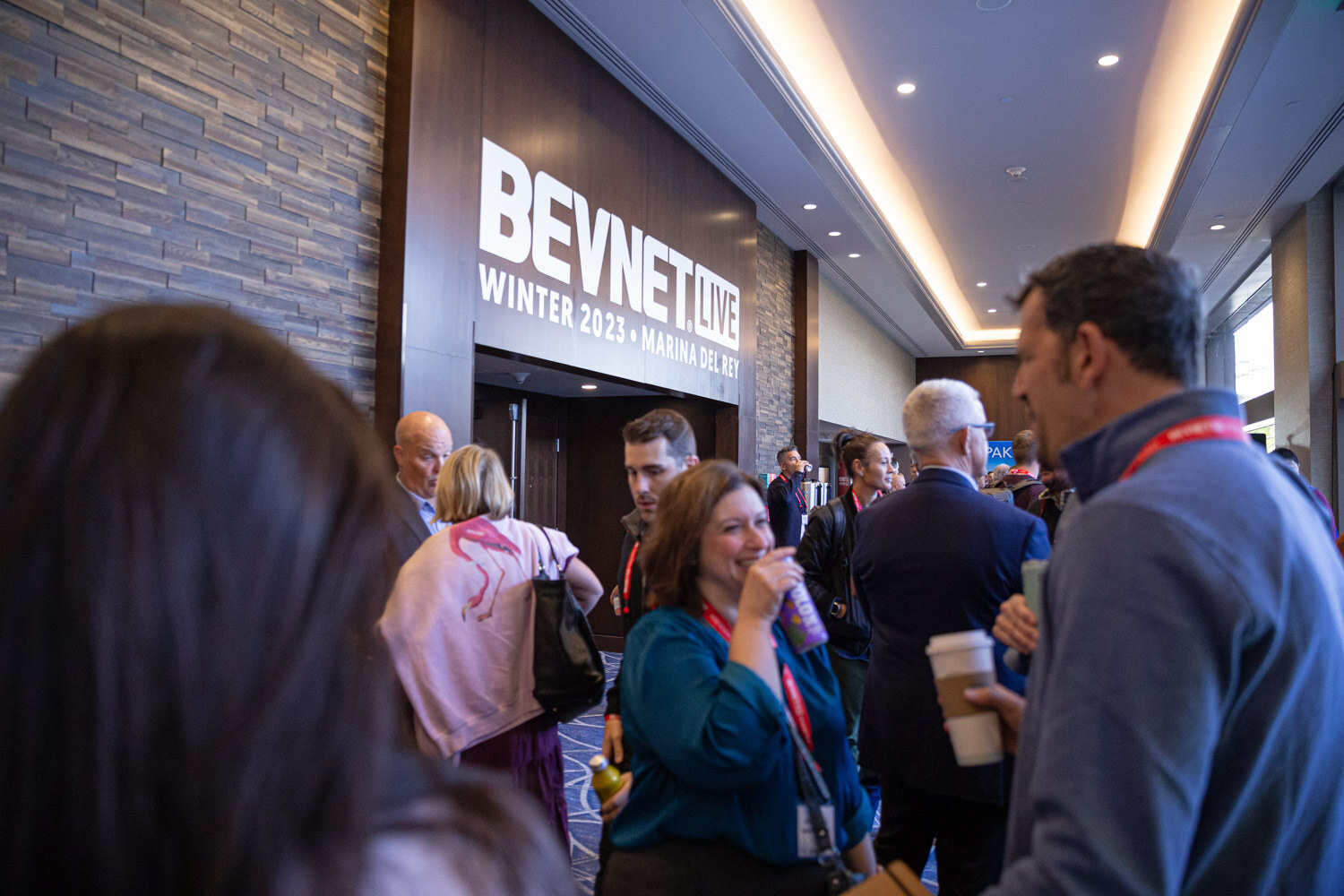 A crowd of attendees stands in front of a wall sign reading BevNET Live Winter 2023 Marina del Rey.