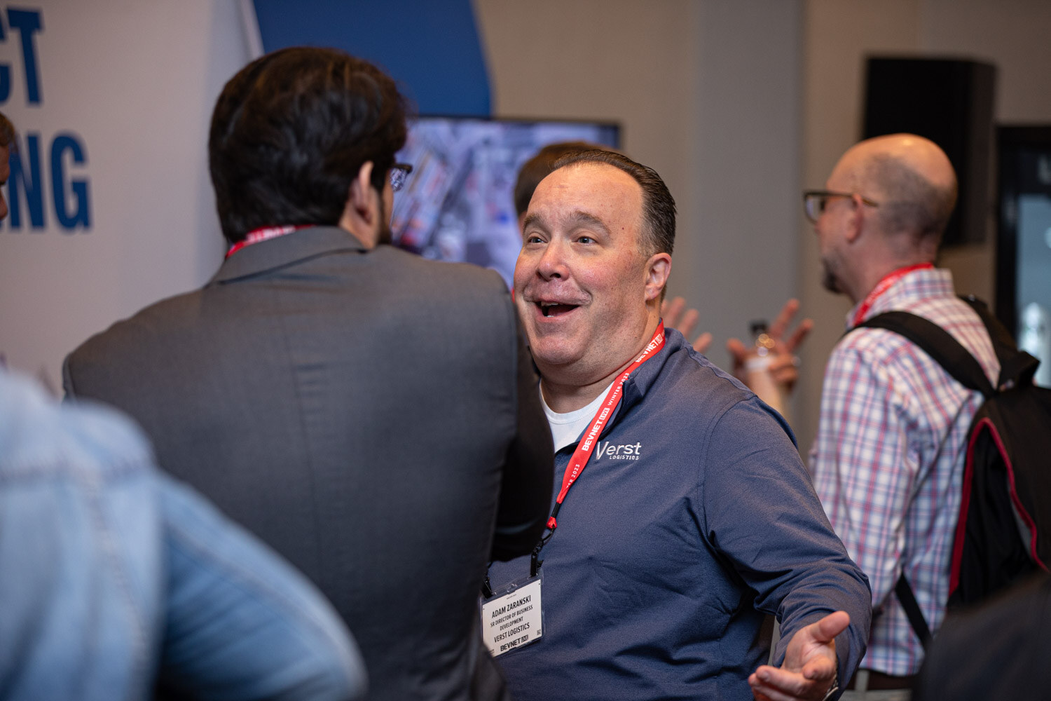 A man in a blue Verst shirt with a lanyard smiles with raised hand while other attendees stand around him.