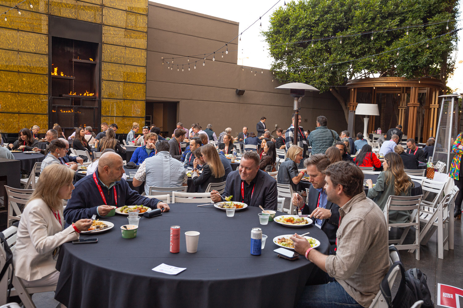 Round tables with black cloths, plates and cups fill a hotel courtyard with attendees seated.