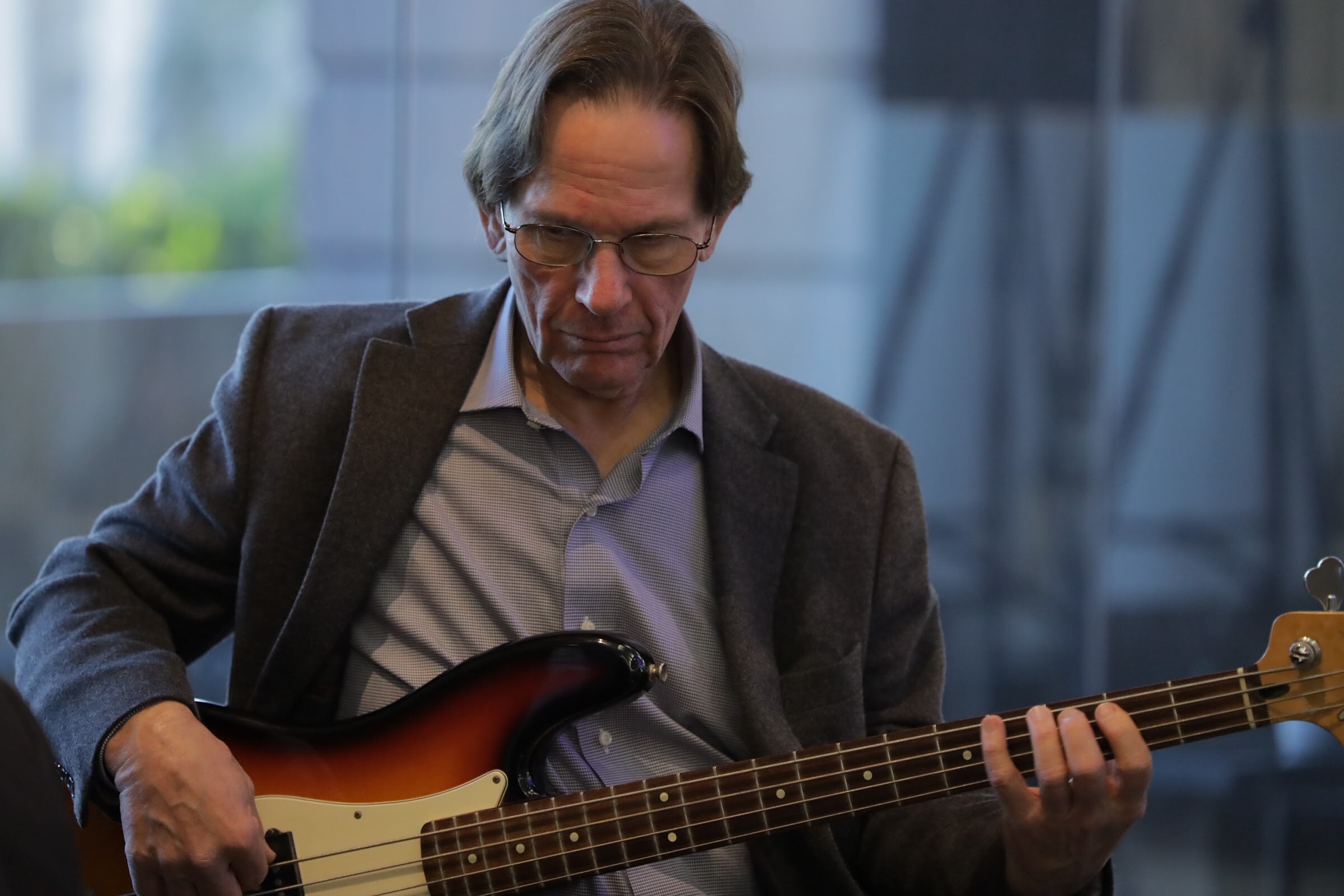 A man with long gray hair and glasses plays an electric bass guitar.