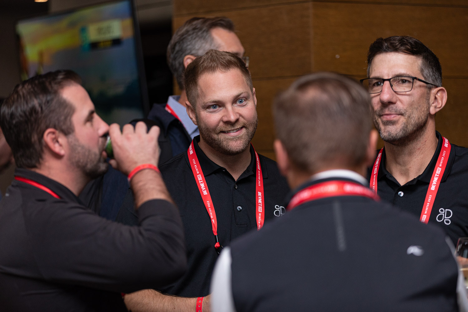 Men in lanyards stand close together; one tilts a can to drink while another smiles.