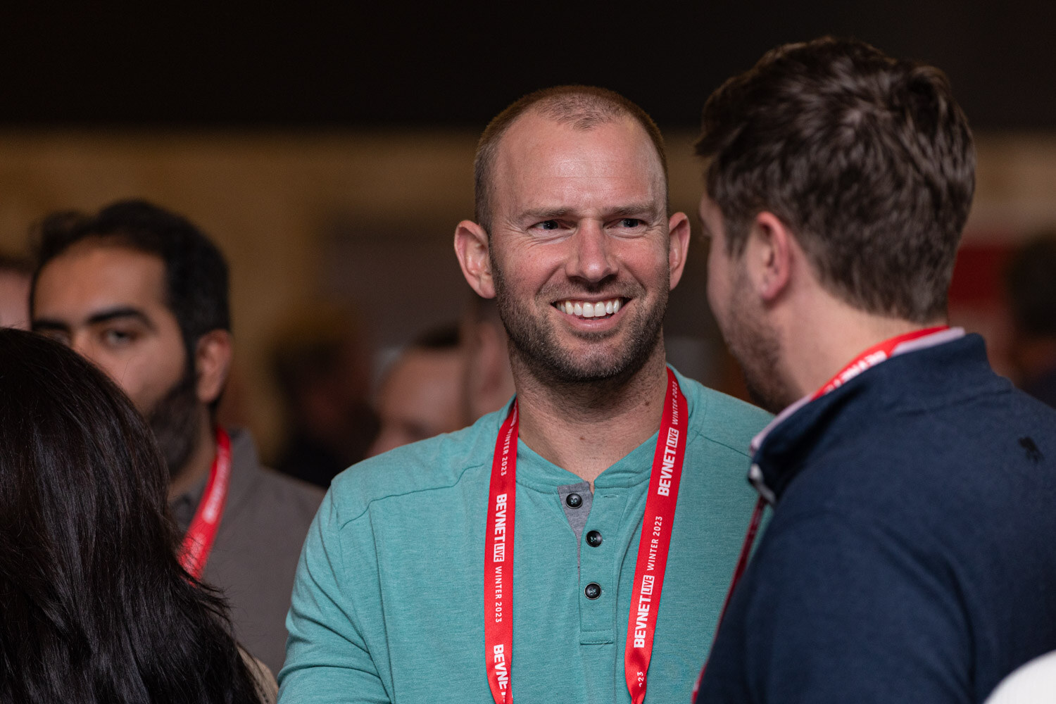 A smiling man with a short beard in a teal shirt stands in front of a crowd.