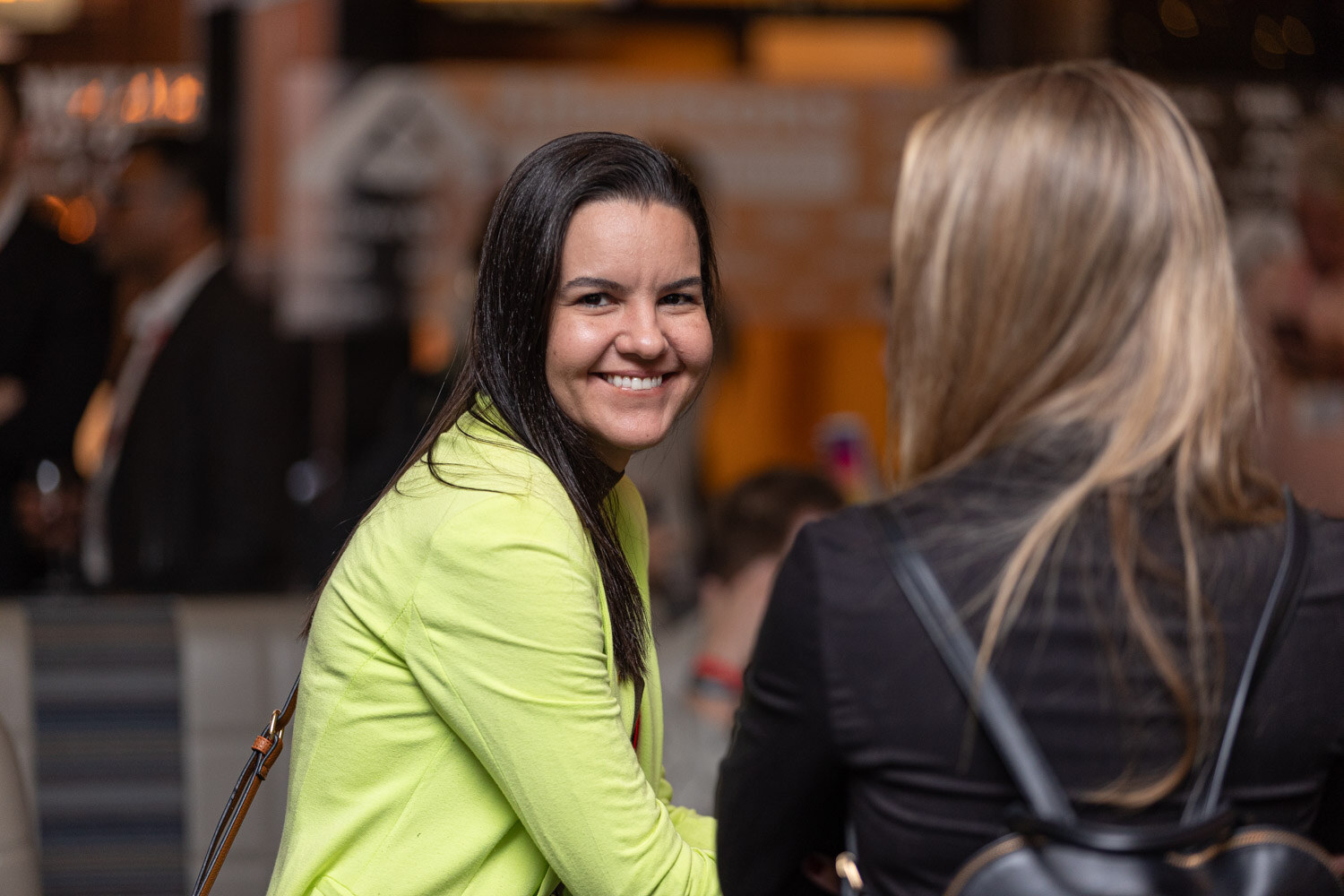 A woman with long dark hair and a lime green blazer smiles while another woman faces away.