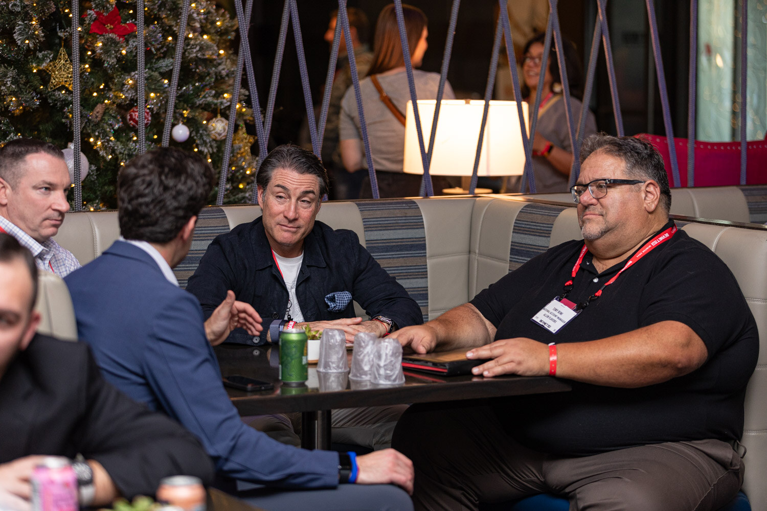 Several men sit at a lounge table with drinks under a decorated Christmas tree.