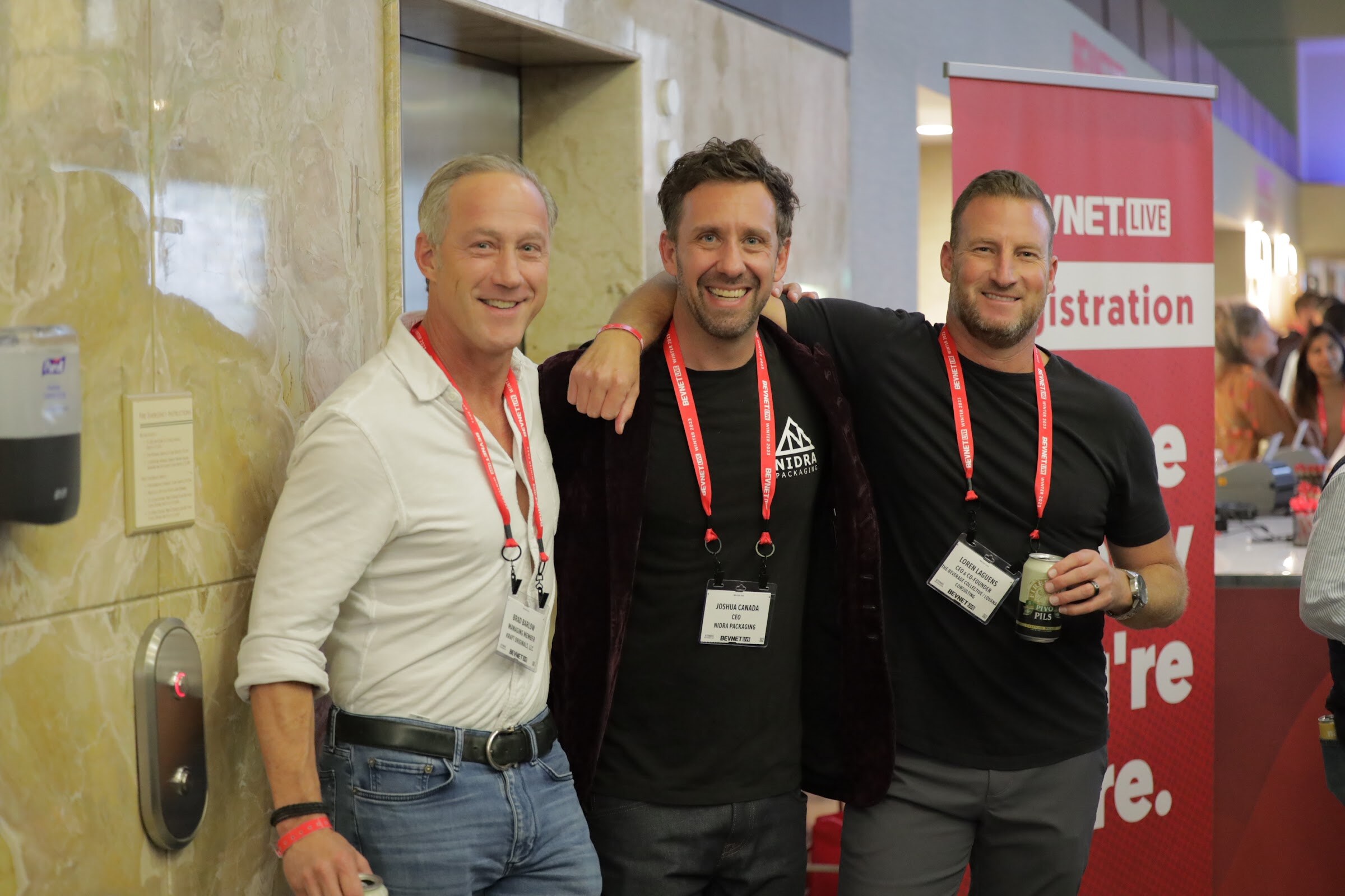 Three men in lanyards smile in front of a red BevNET Live registration banner.