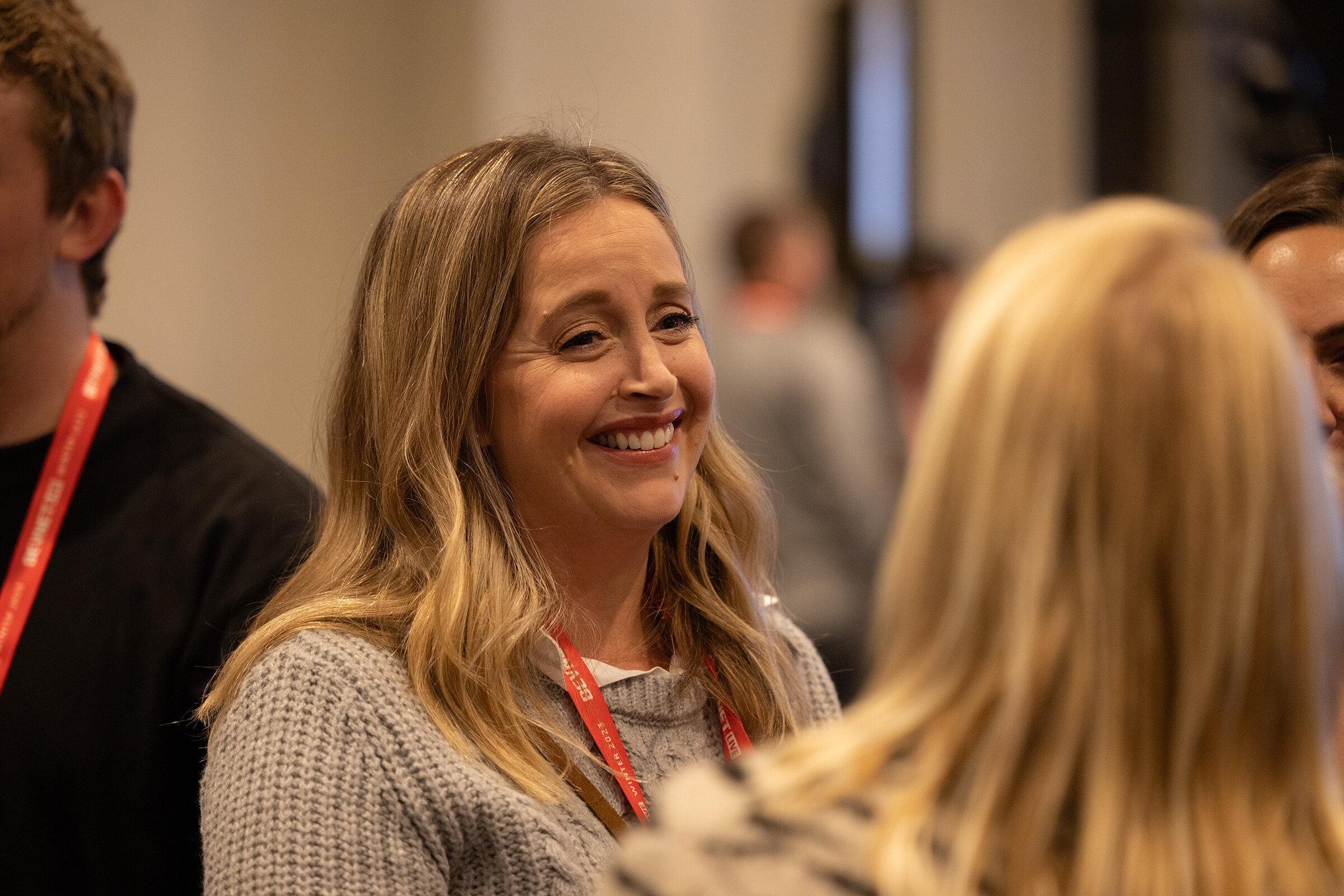 A woman with long blond hair and a red lanyard smiles while talking with another attendee.