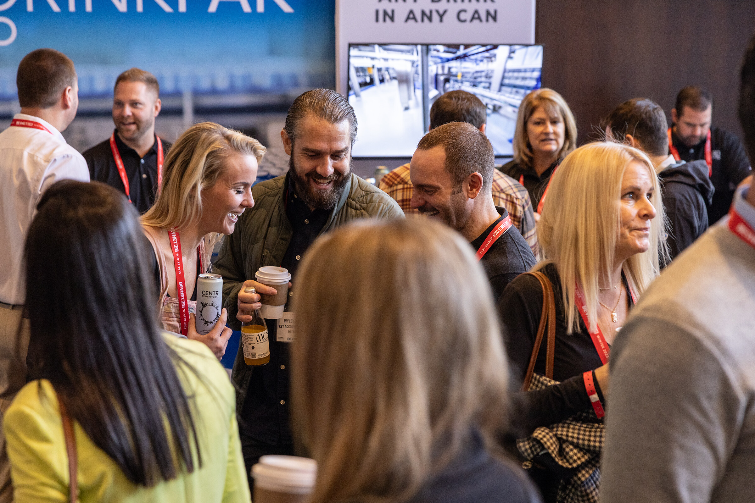 A group of attendees in lanyards laugh together, holding cups and beverage cans.