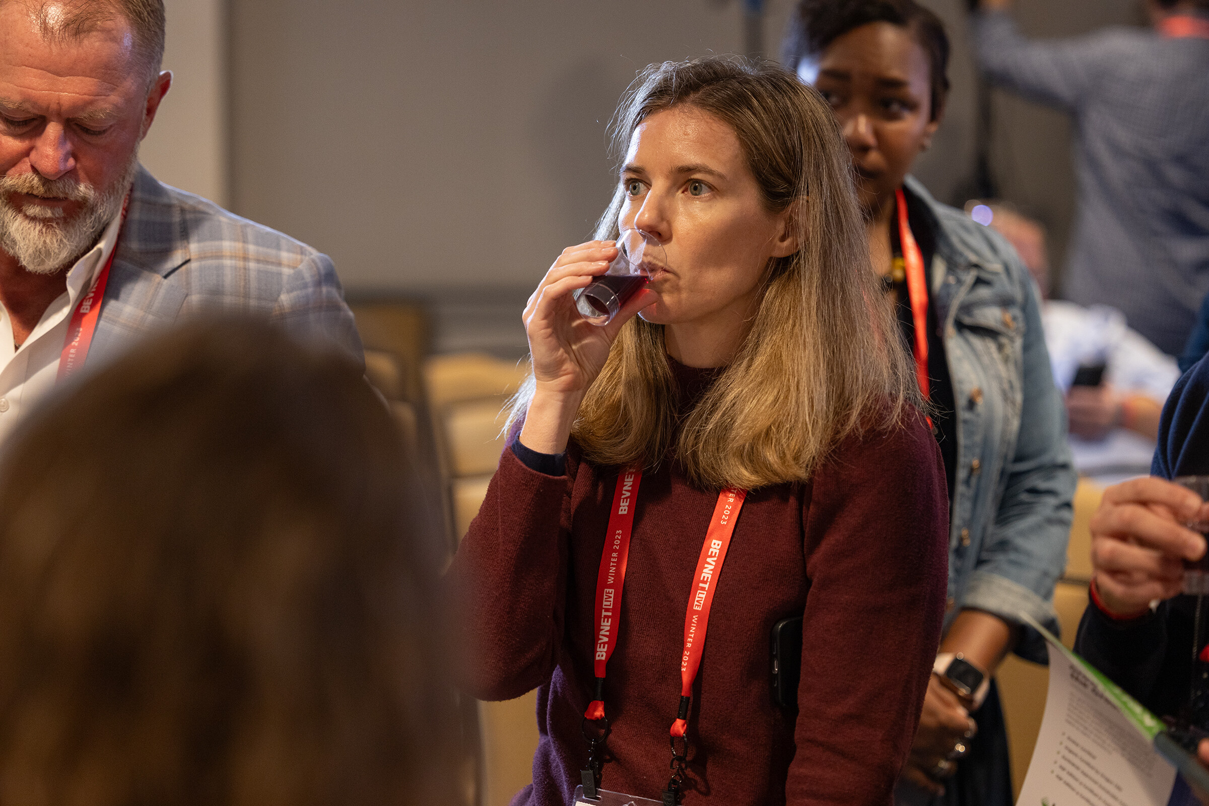 A woman in a maroon sweater drinks from a small clear cup while other attendees stand around her.