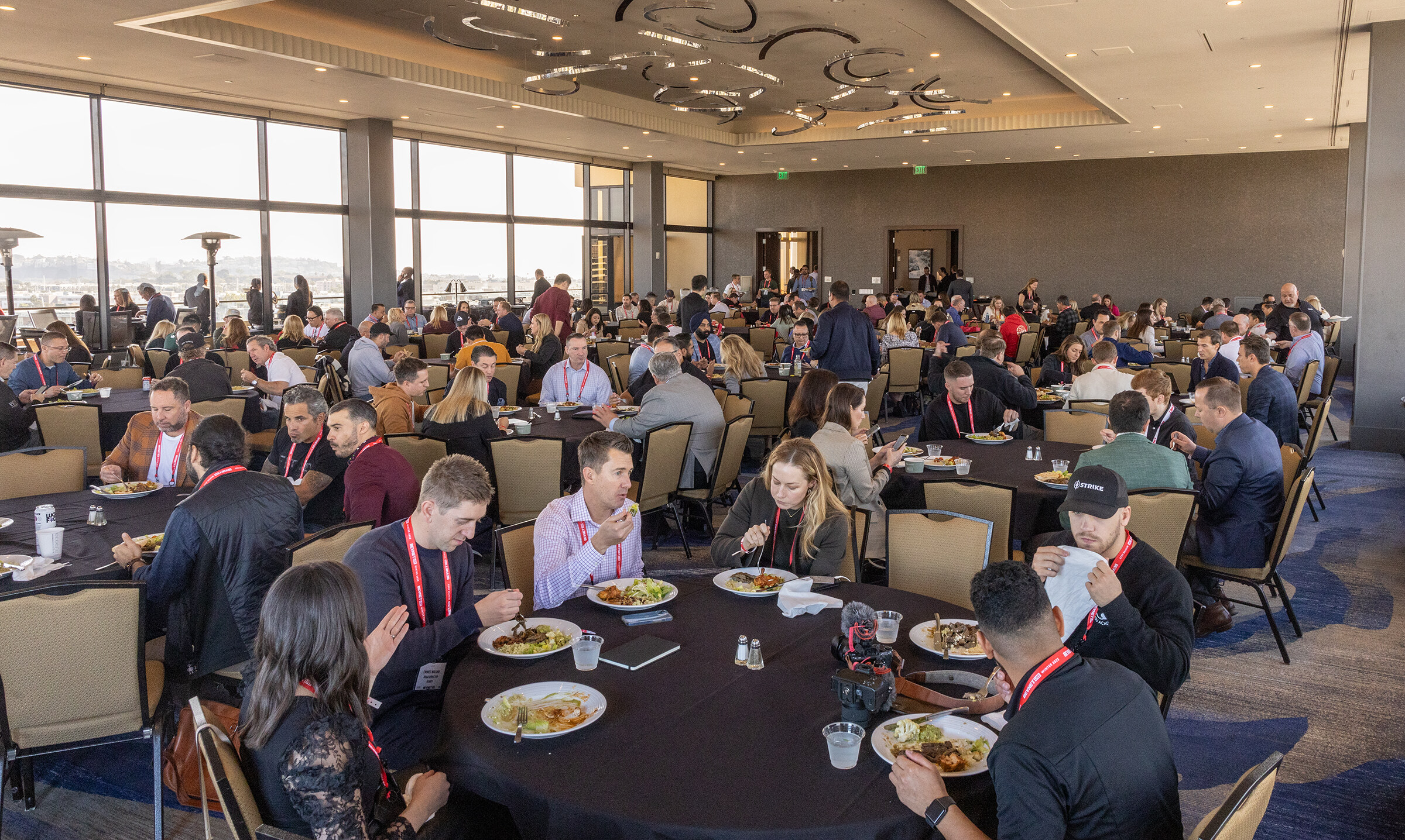 A wide view of attendees seated at round tables with plates of food in a ballroom with window views.