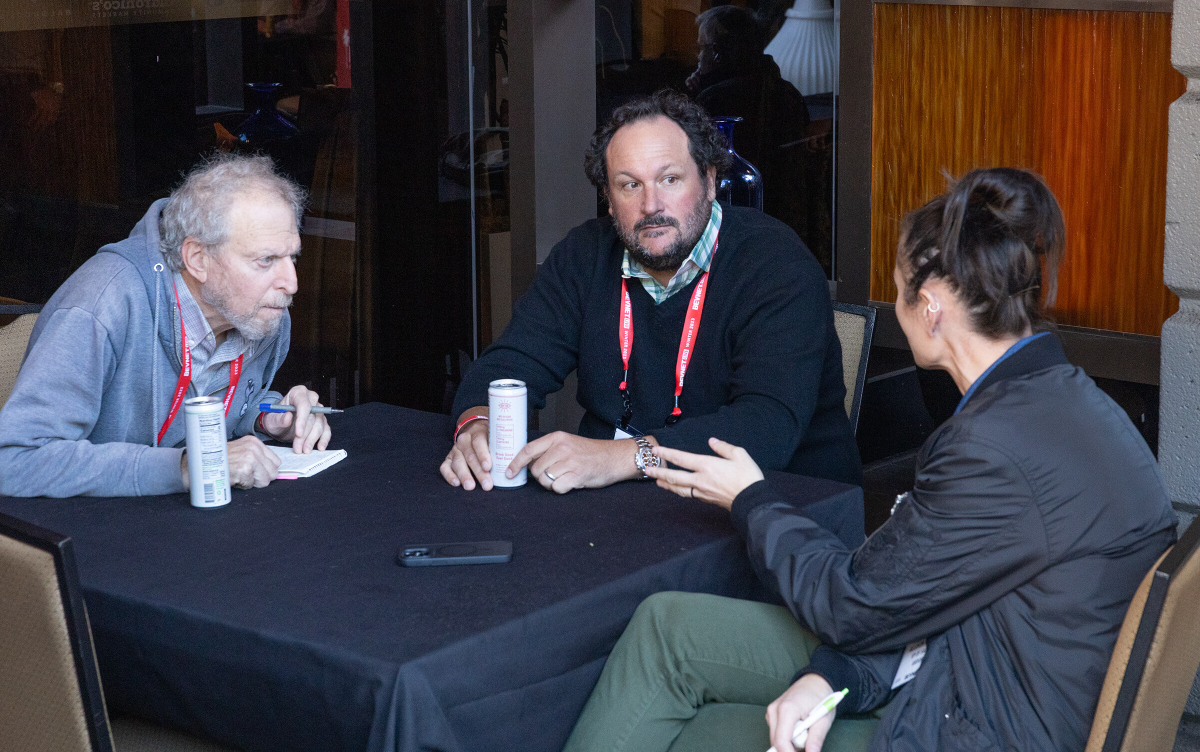 Three people with lanyards sit around a small table with a beverage can and notes.