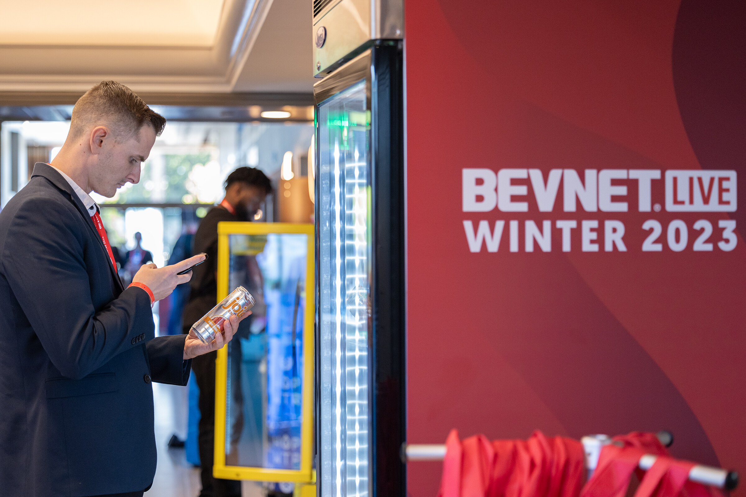 A man in a dark suit looks at a phone next to an open drink cooler and a red BevNET Live Winter 2023 banner.