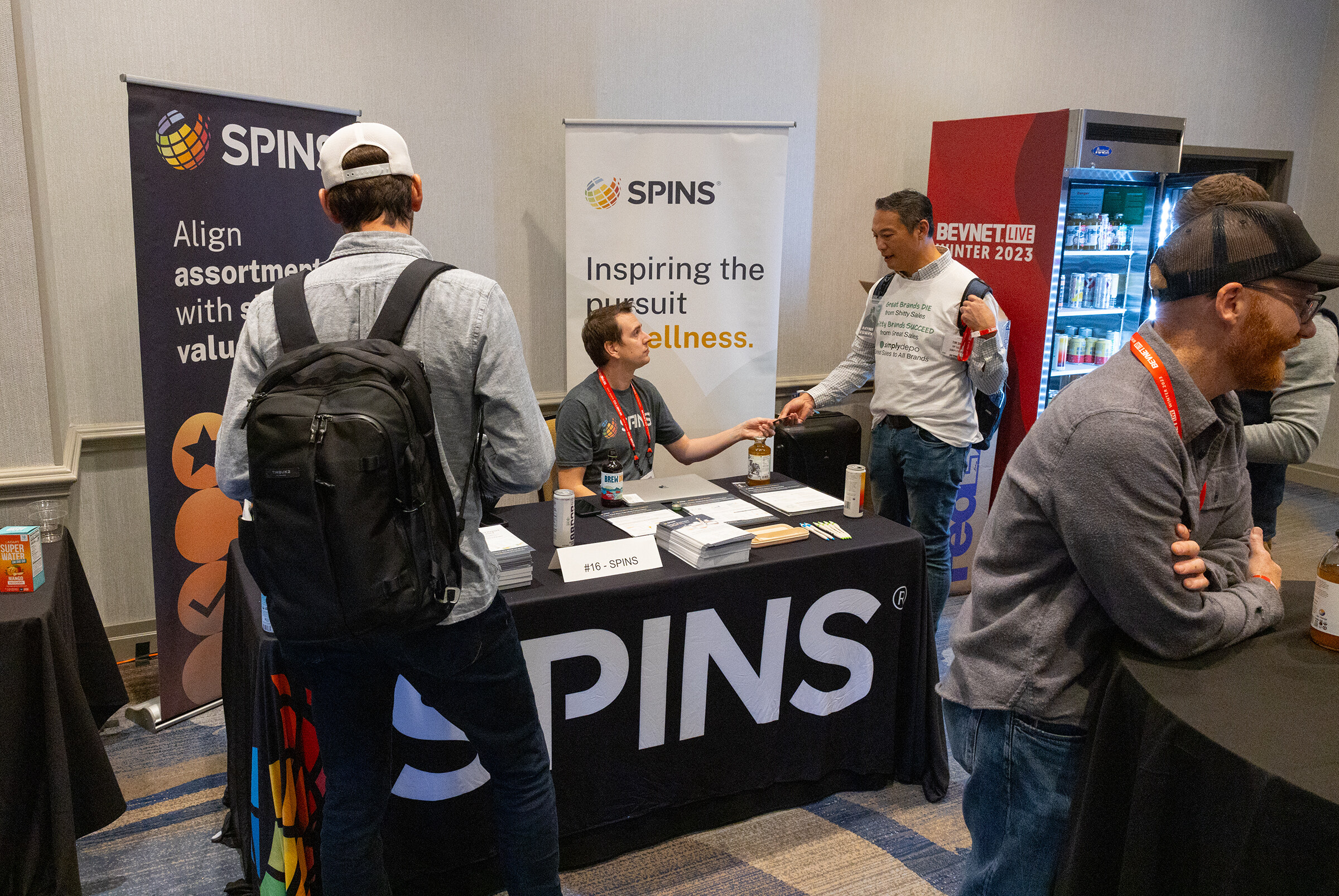 People talk across a black-draped table with SPINS banners and a refrigerator behind them.