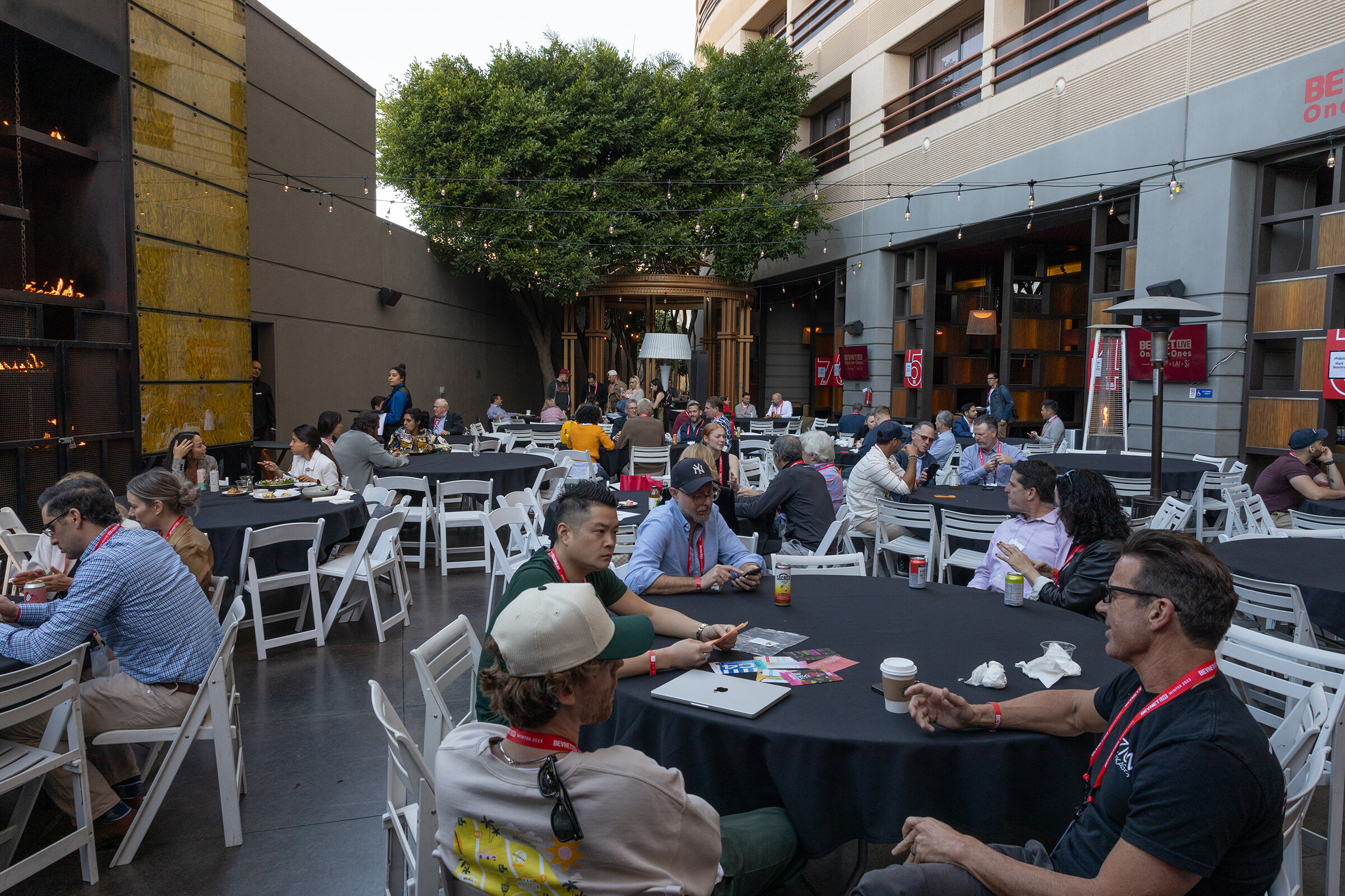 Attendees sit at round tables in a sunny courtyard surrounded by hotel buildings and a tree.