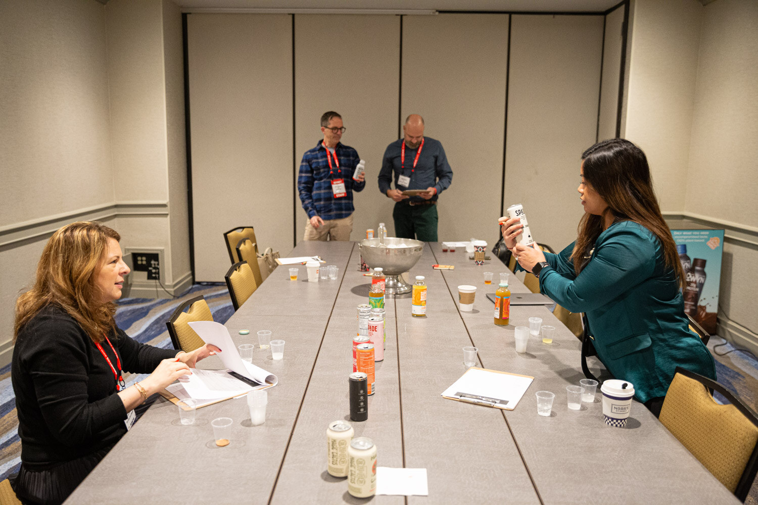 Judges sit and stand around a long table with samples in small cups, in a meeting room.