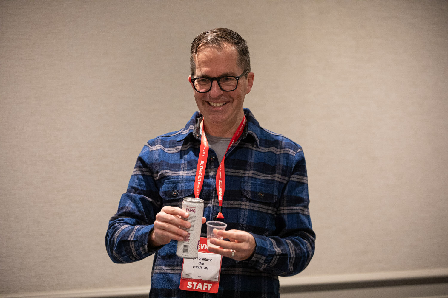 A smiling man in a plaid shirt and red staff lanyard holds a beverage can and cup.