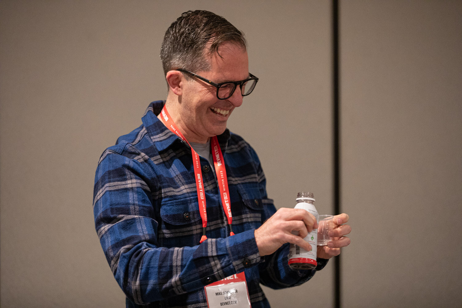 A man in a plaid shirt and staff lanyard laughs while holding a bottle and cup.