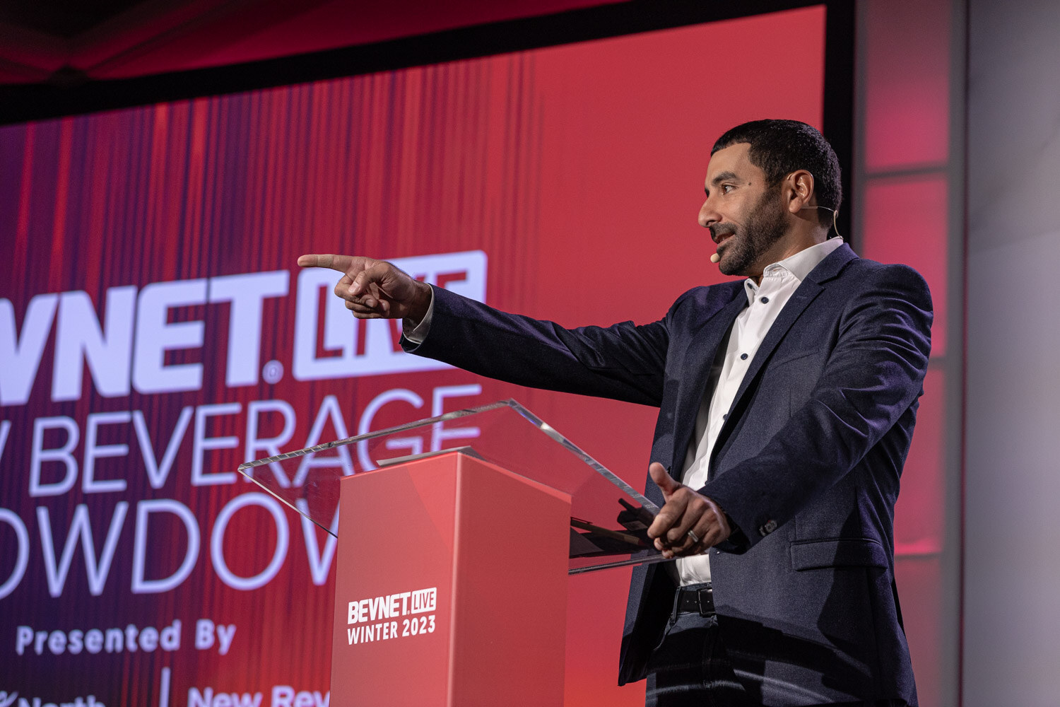 A man in a navy blazer points while standing at a red lectern in front of a red New Beverage Showdown screen.