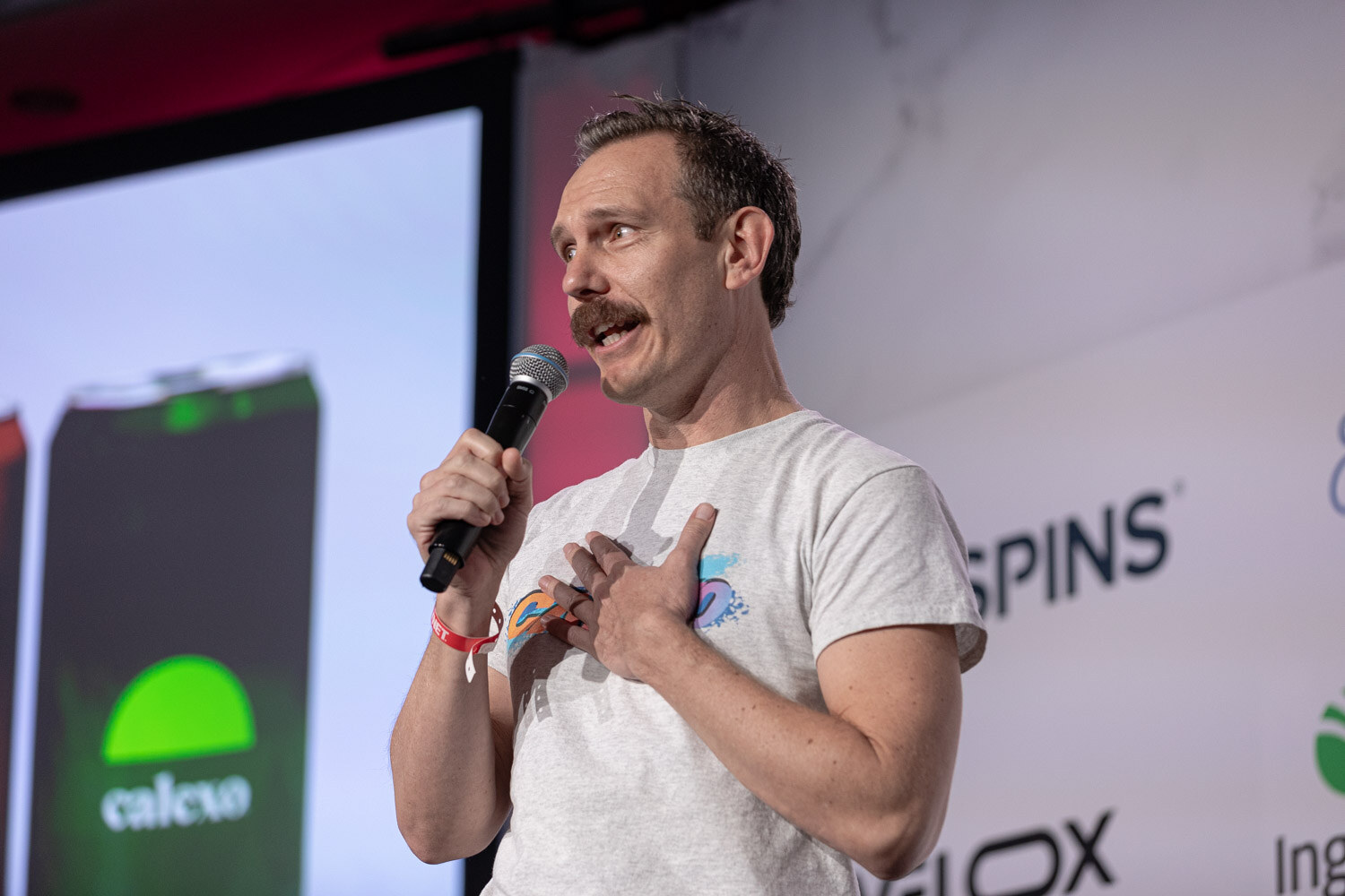 A man in a gray T-shirt speaks into a microphone in front of a slide with colorful cans.