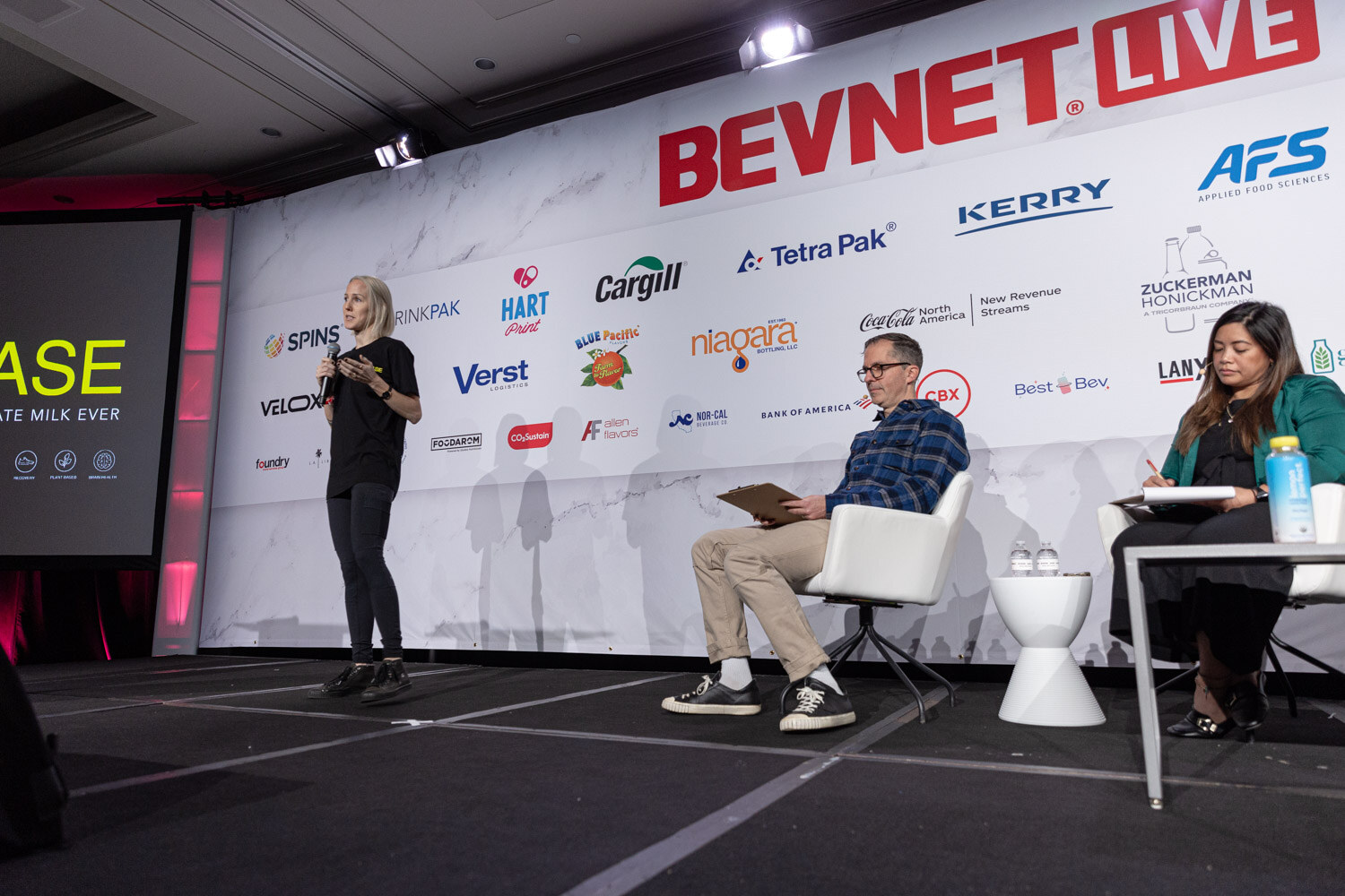A woman stands on stage left while two judges and a host sit on stage right in front of a sponsor wall.
