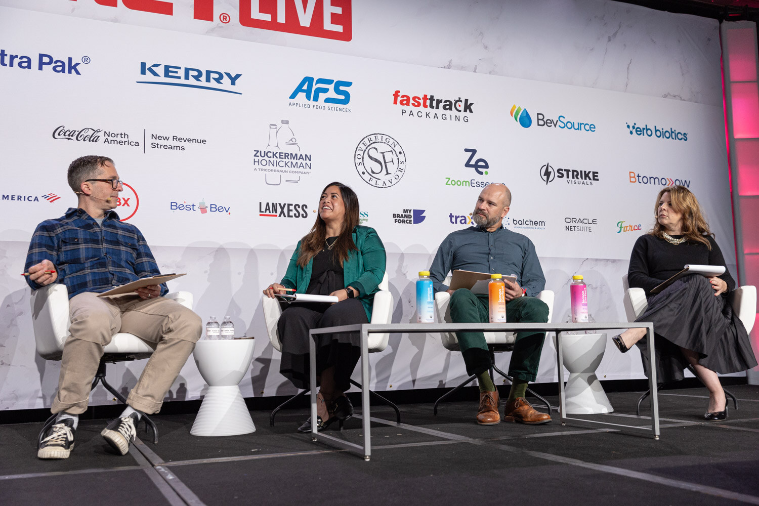 Four people sit on stage on white stools, with a sponsor wall behind them.