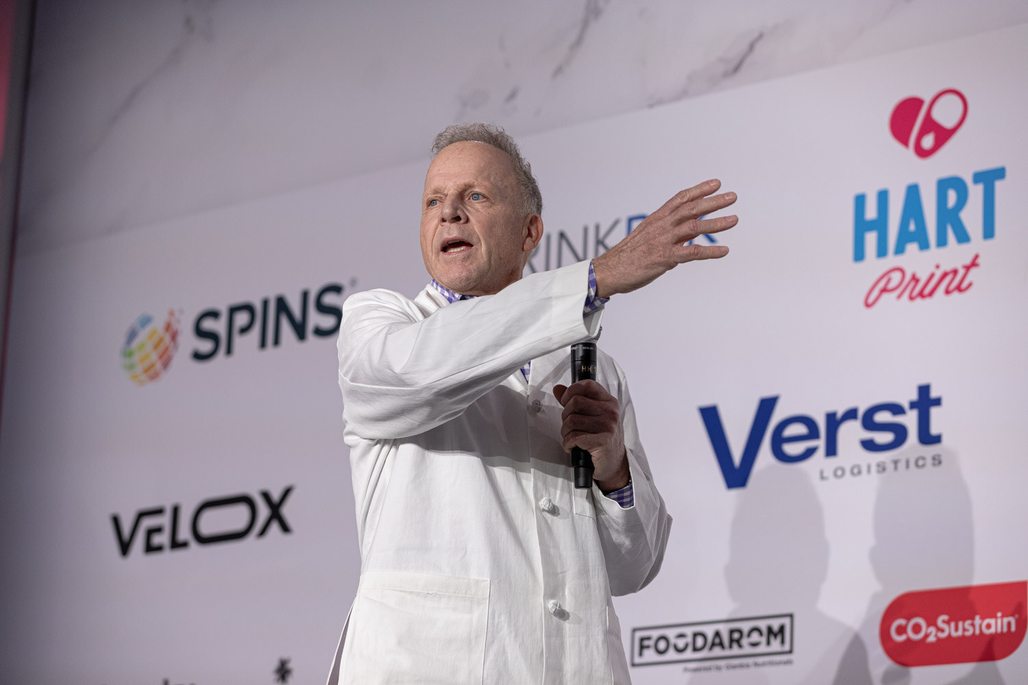 A man in a white lab coat extends one arm while speaking into a microphone.