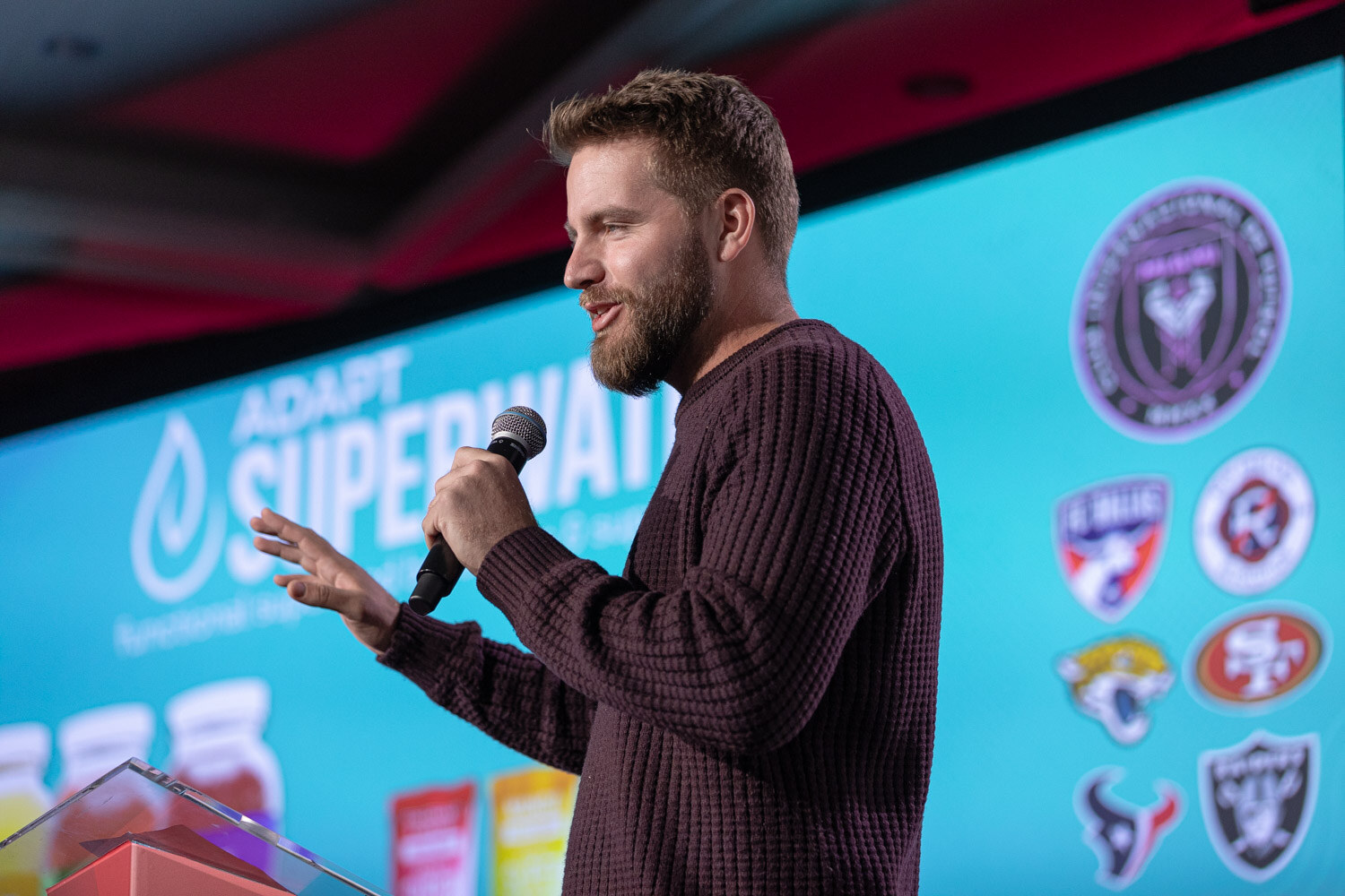 A bearded man in a maroon sweater speaks in front of a blue slide that reads Adapt Superwater.