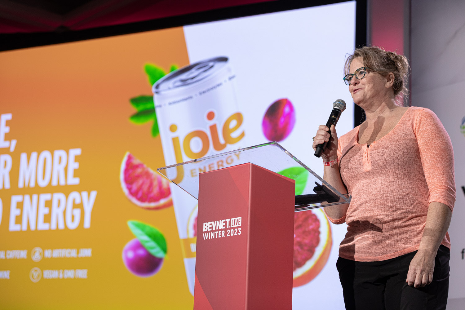 A woman in an orange blouse and glasses speaks at a red lectern with a Joie slide behind her.