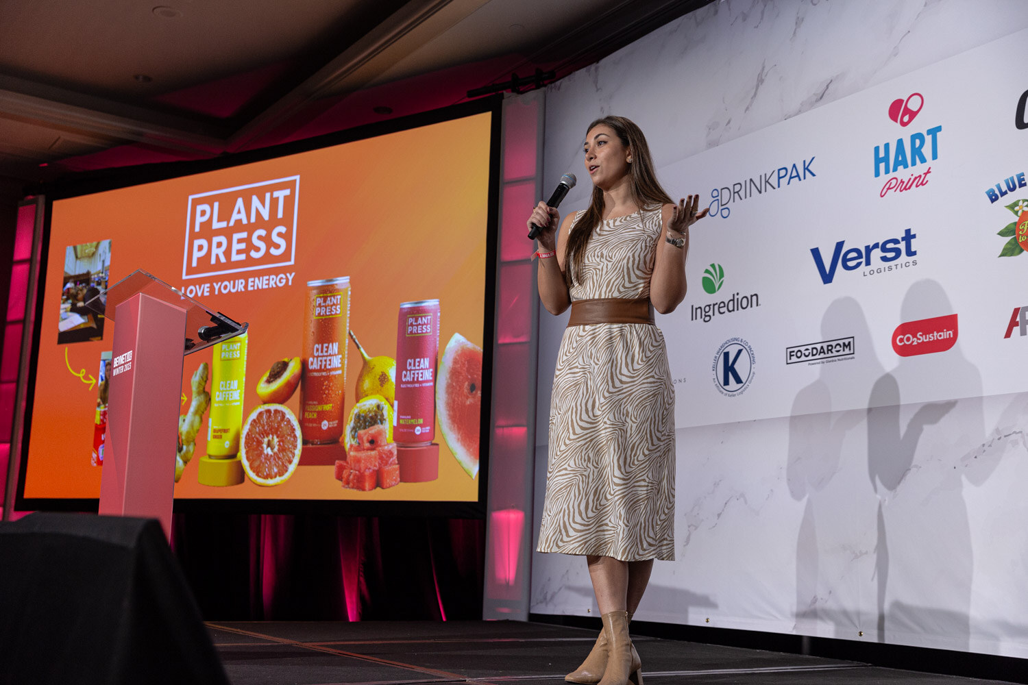 A woman in a patterned dress stands on a stage beside a slide that reads Plant Press.