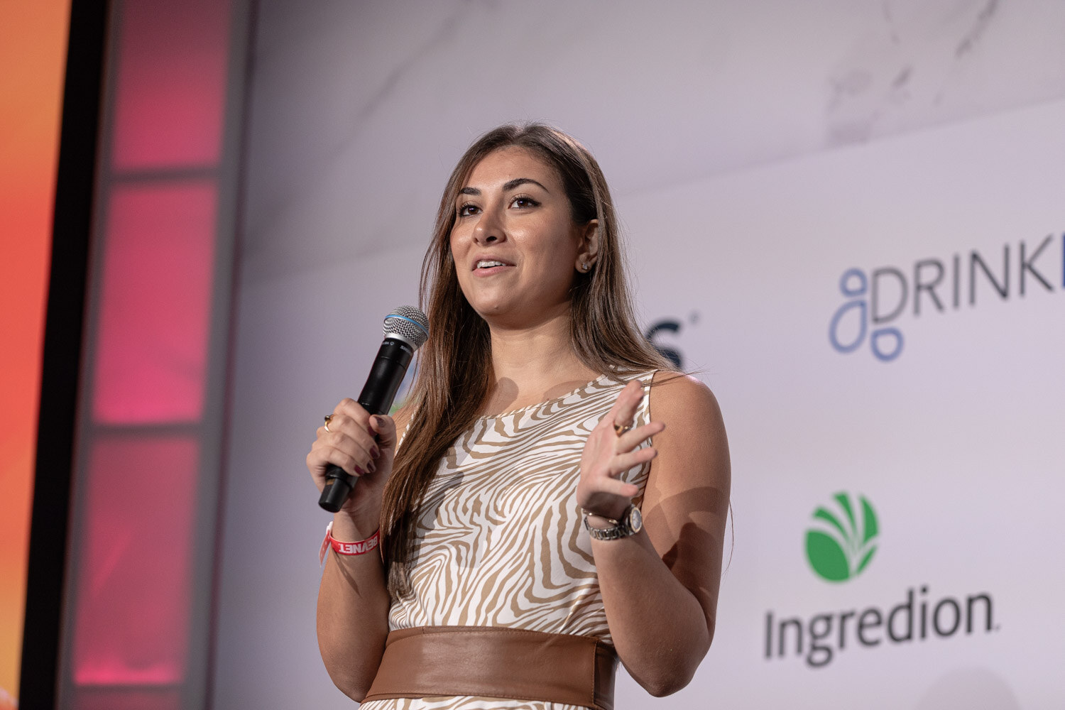 A woman in a patterned dress and brown belt speaks into a microphone in front of an orange wall.