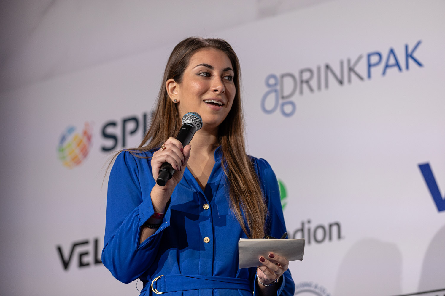 A woman in a blue dress holds a microphone and index cards in front of a sponsor wall.