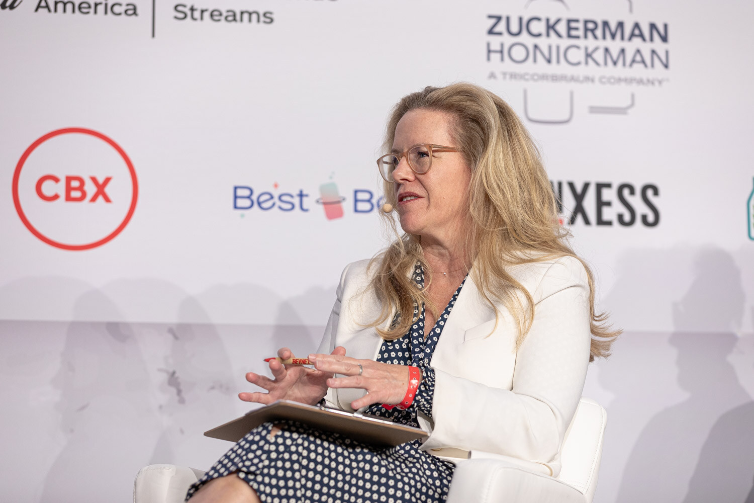 A woman in a white blazer with long blond hair speaks while seated, holding a clipboard.