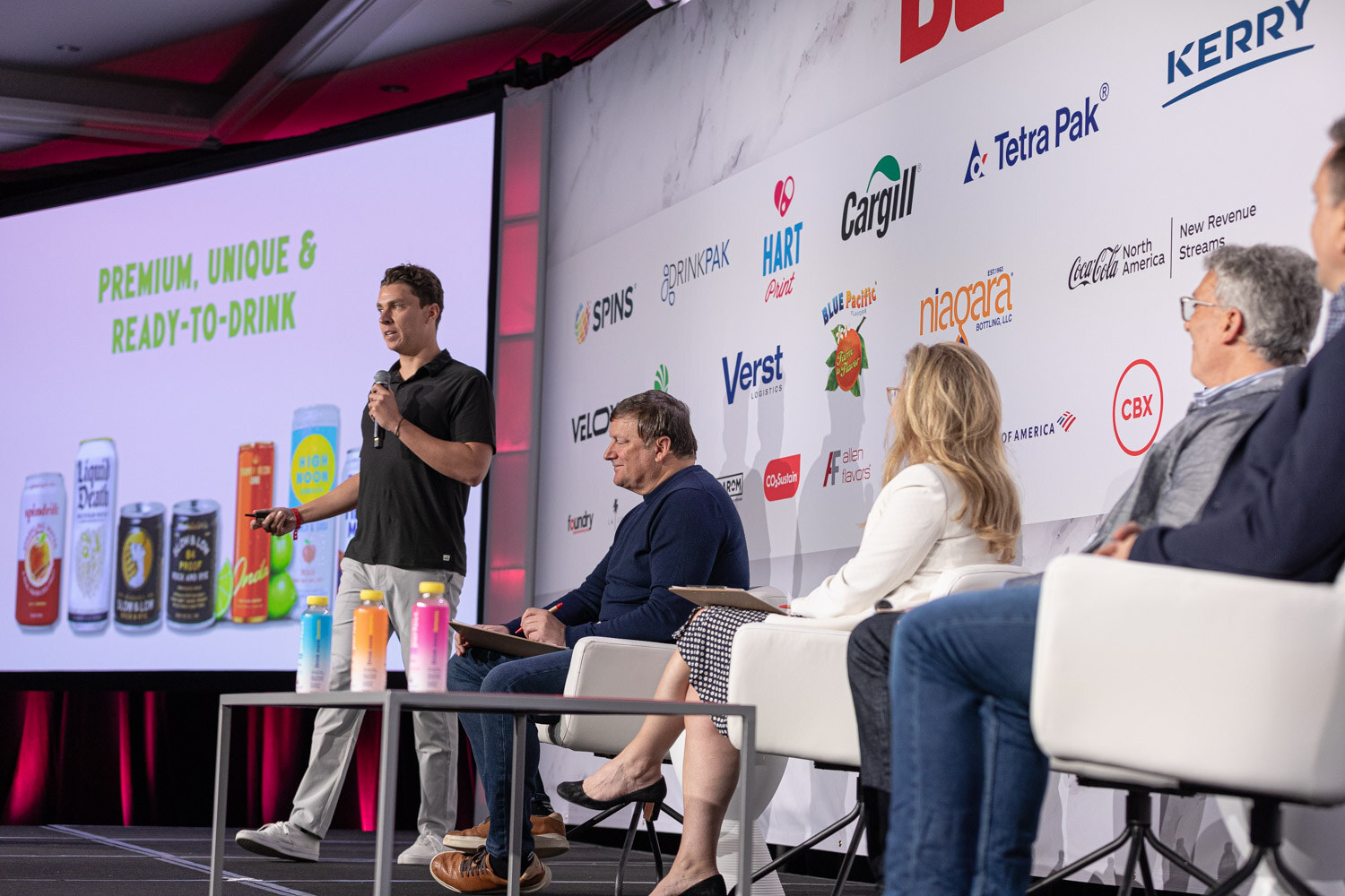 A man in a black polo stands beside a panel of judges seated on stage in front of a slide of cans.