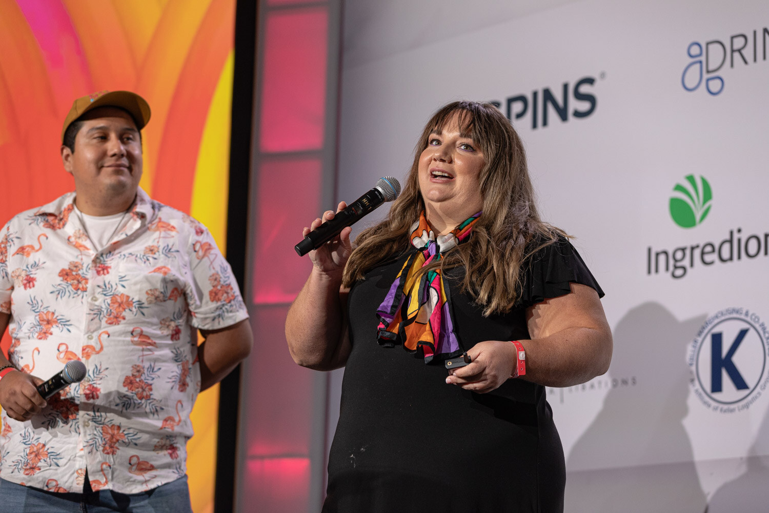 A man in a floral shirt and cap and a woman in a black top hold microphones in front of a colorful slide.
