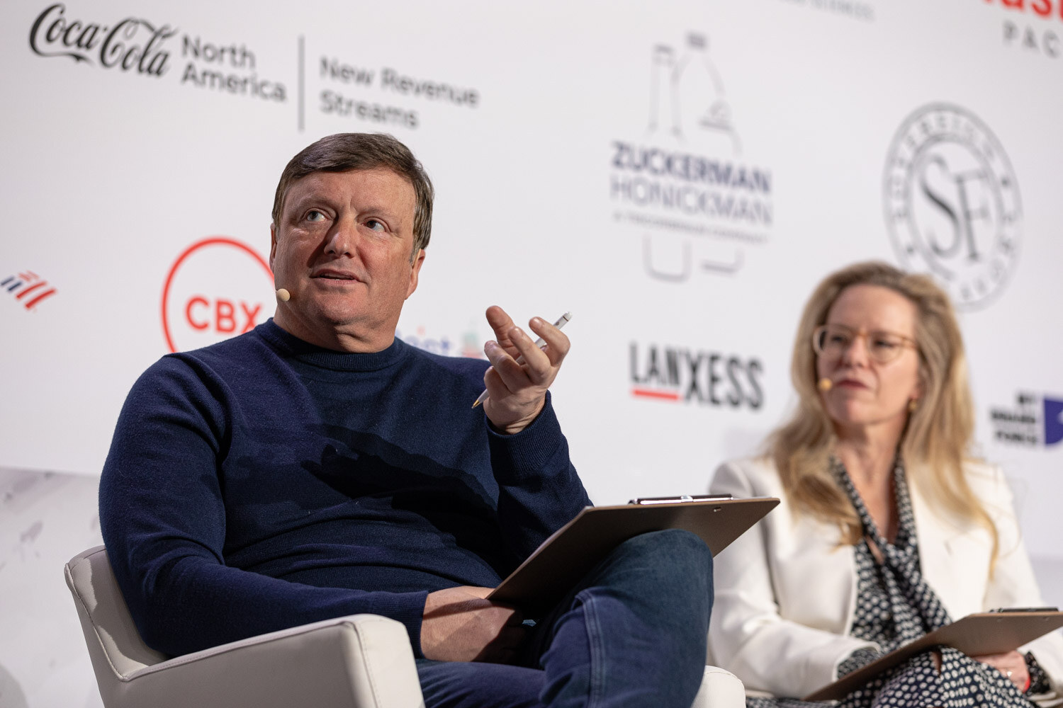 A man in a navy sweater holding a clipboard and a woman in a white blazer sit on stage.