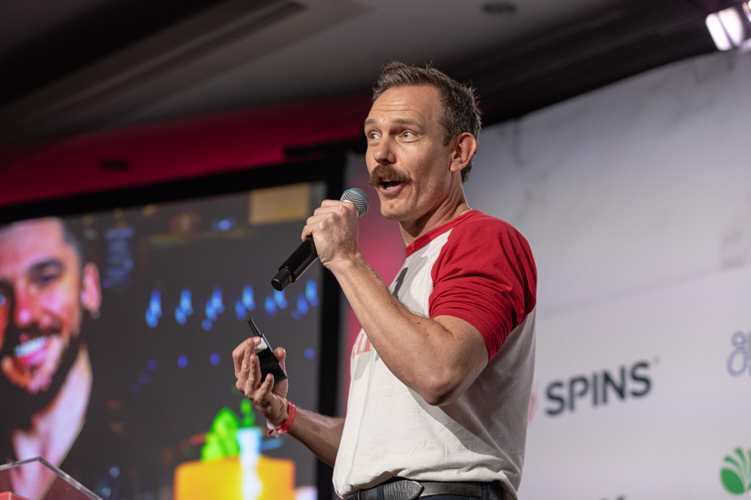 A man with a mustache wearing a red and white raglan shirt speaks into a microphone in front of a screen.