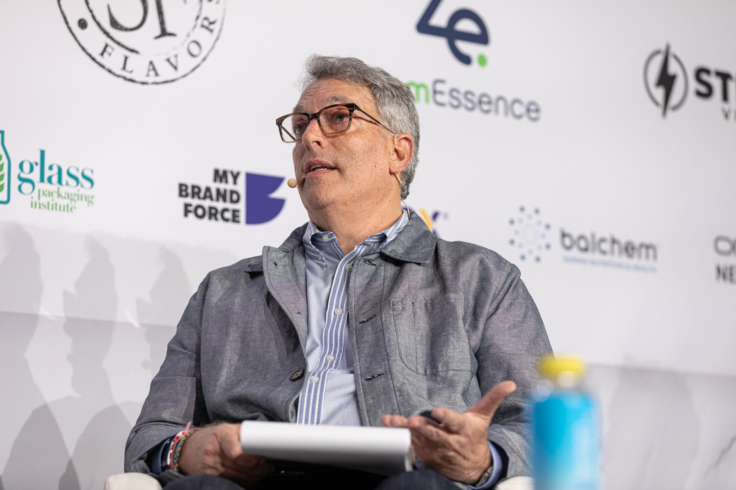 A man in a gray jacket with glasses looks off-stage while seated, holding notes.