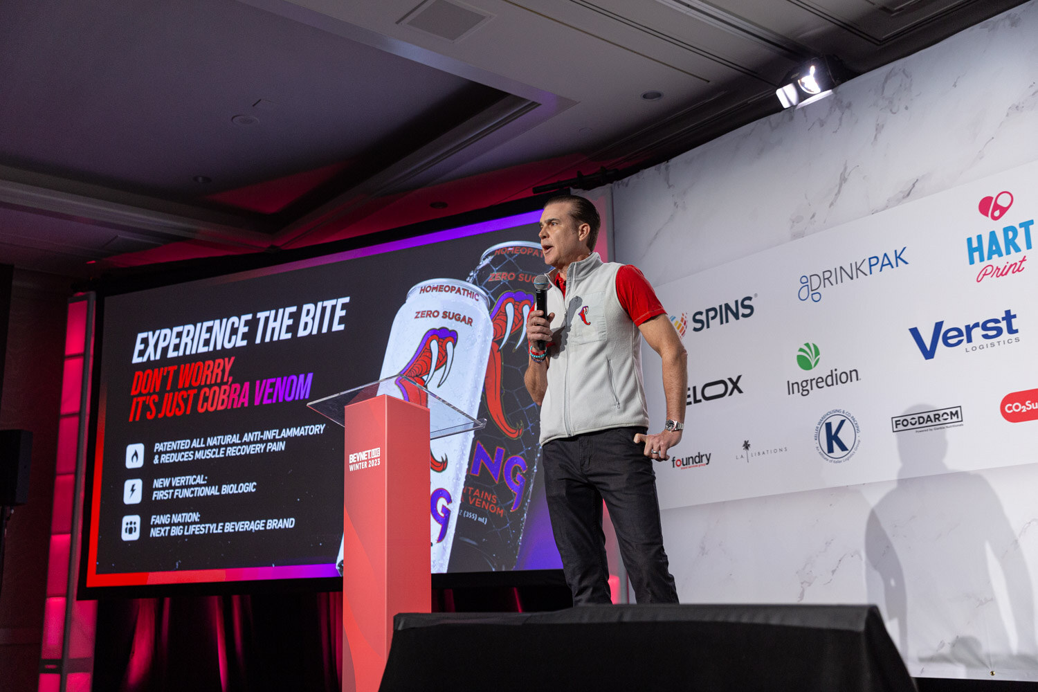 A man in a gray vest stands at a red lectern next to a screen that reads Experience the bite.