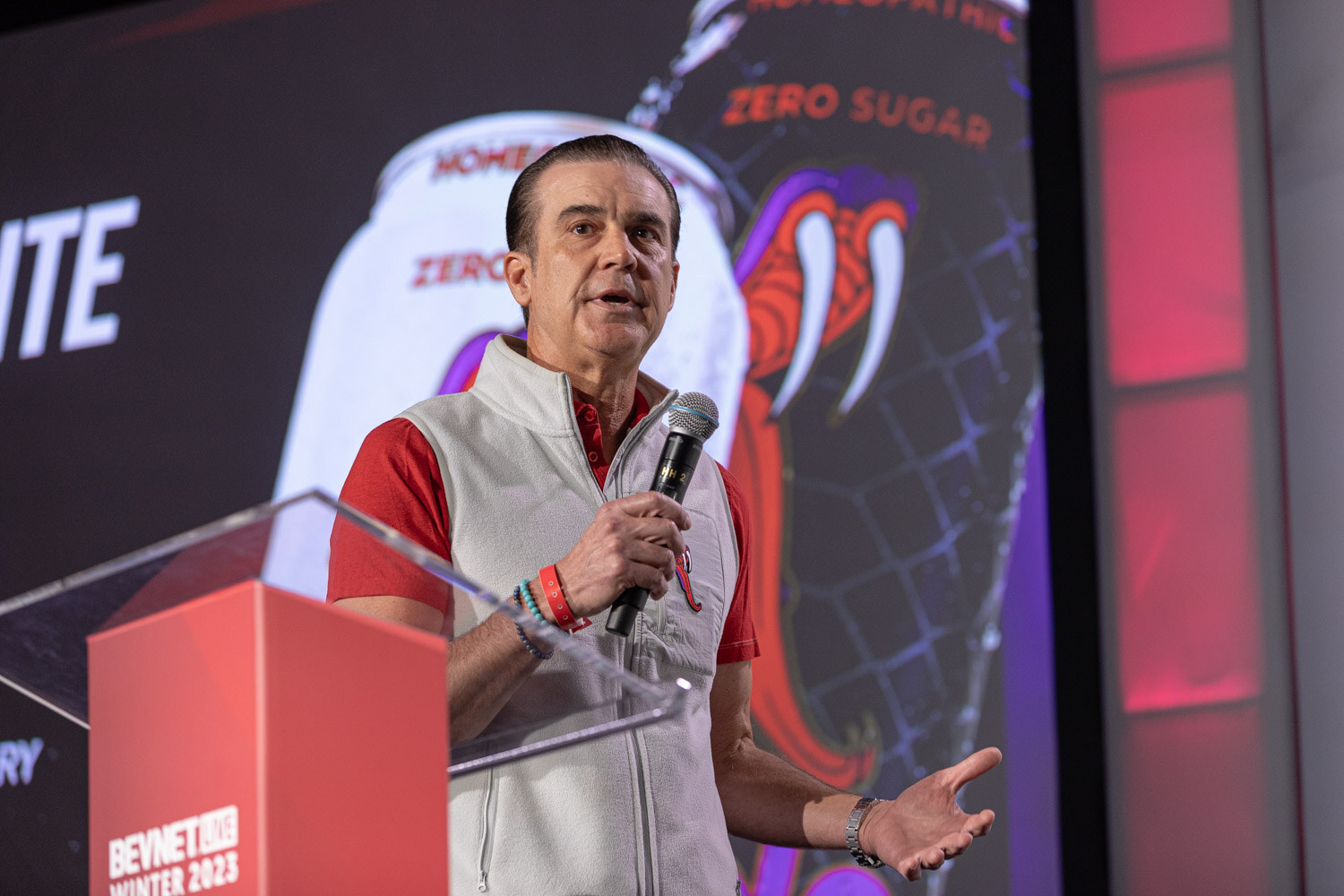 A man in a gray vest speaks into a microphone in front of a slide showing a can with a snake graphic.