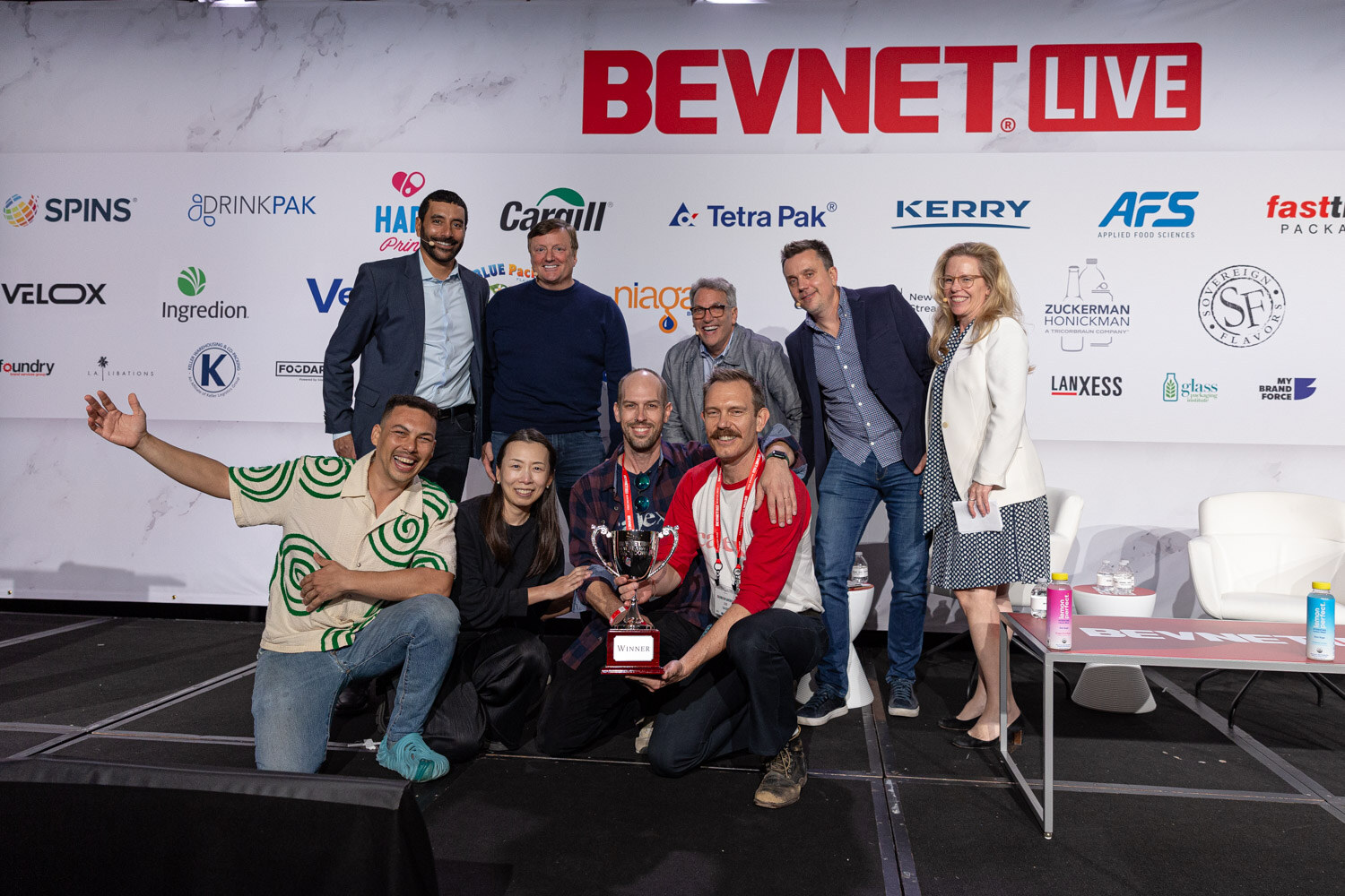A large group of people pose on a stage in front of a BevNET Live backdrop, holding a trophy.