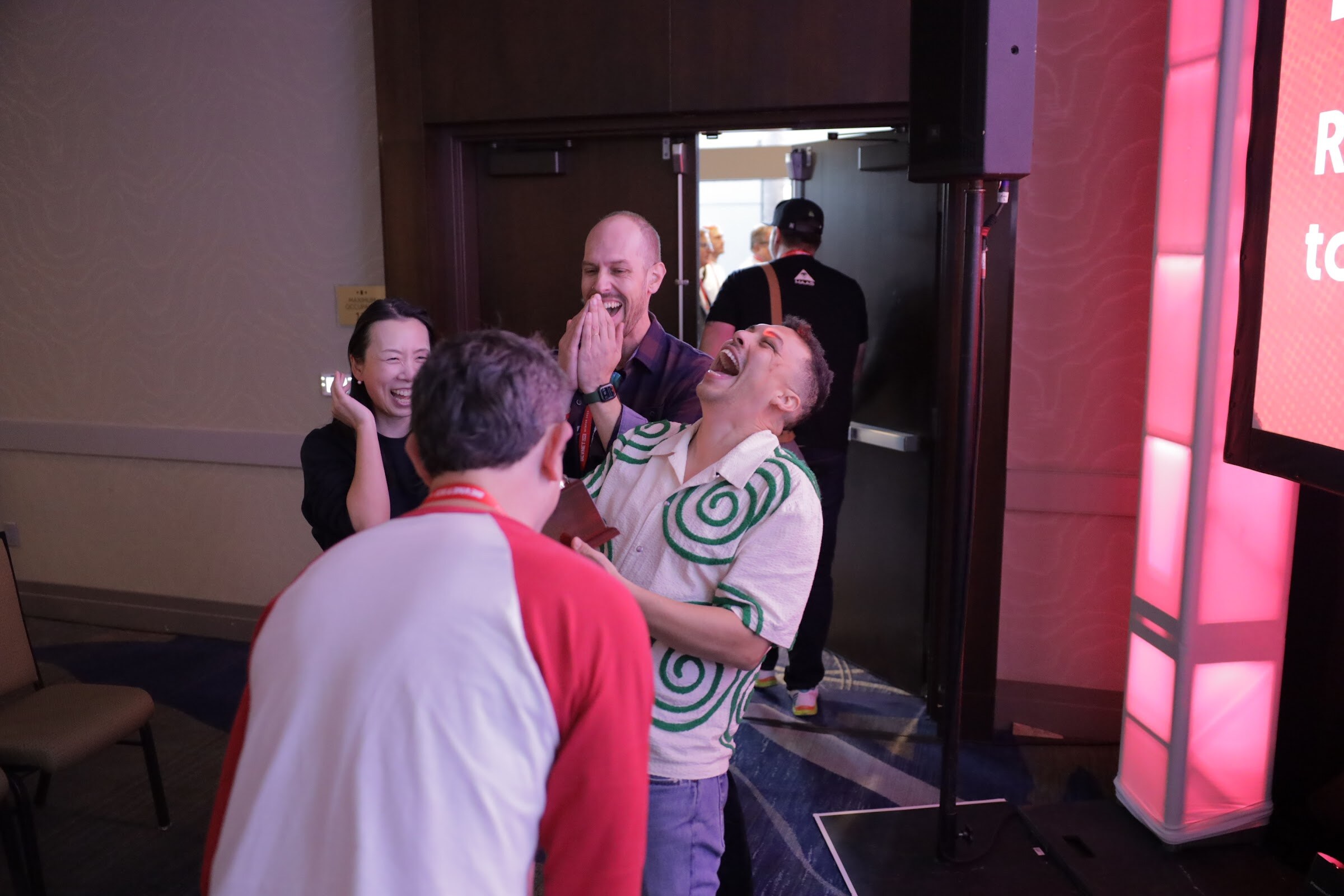 A group of people laugh and hug beside a lit pink panel; a man in a red and white shirt turns away.