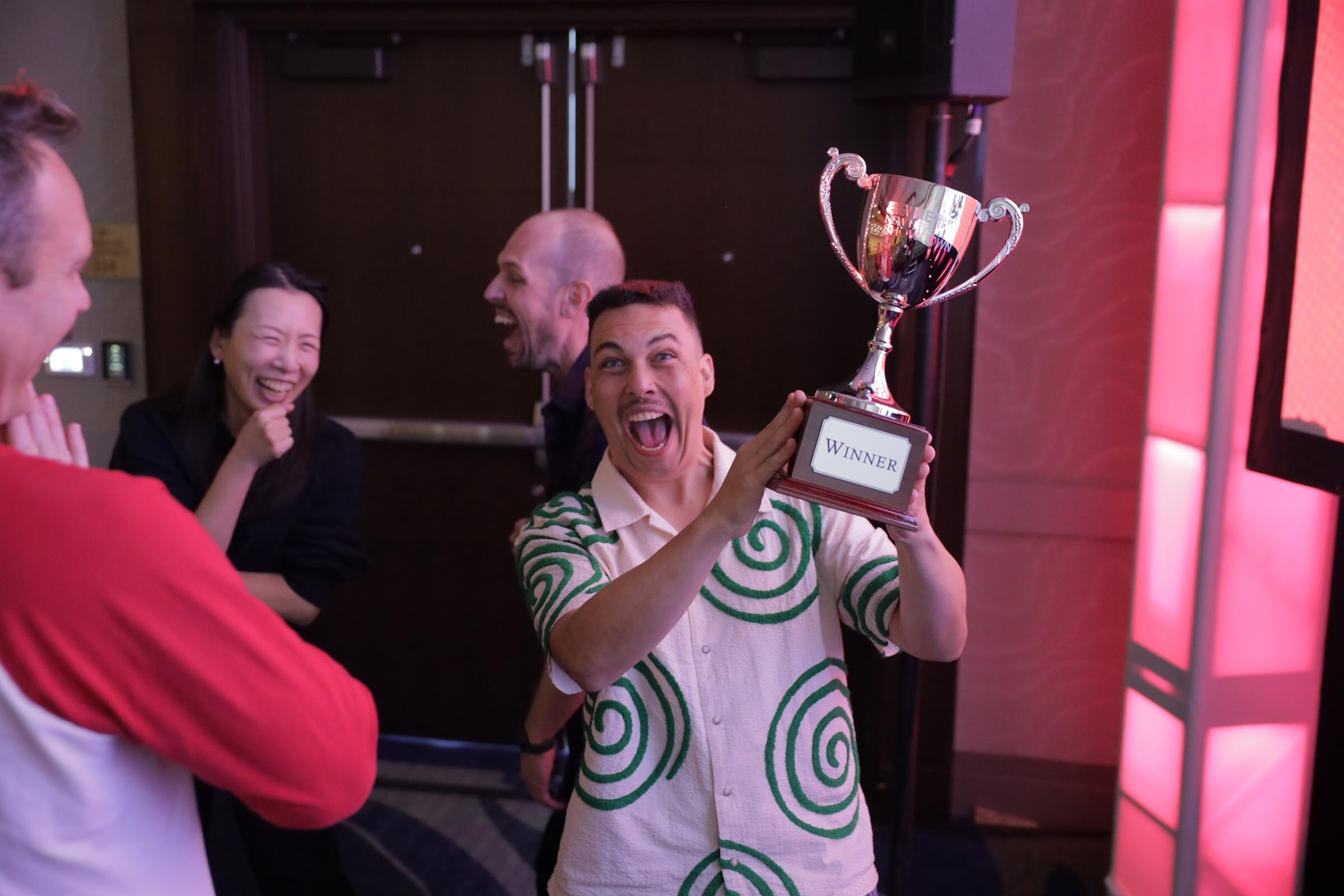 A man in a green and white patterned shirt raises a silver trophy while others laugh around him.