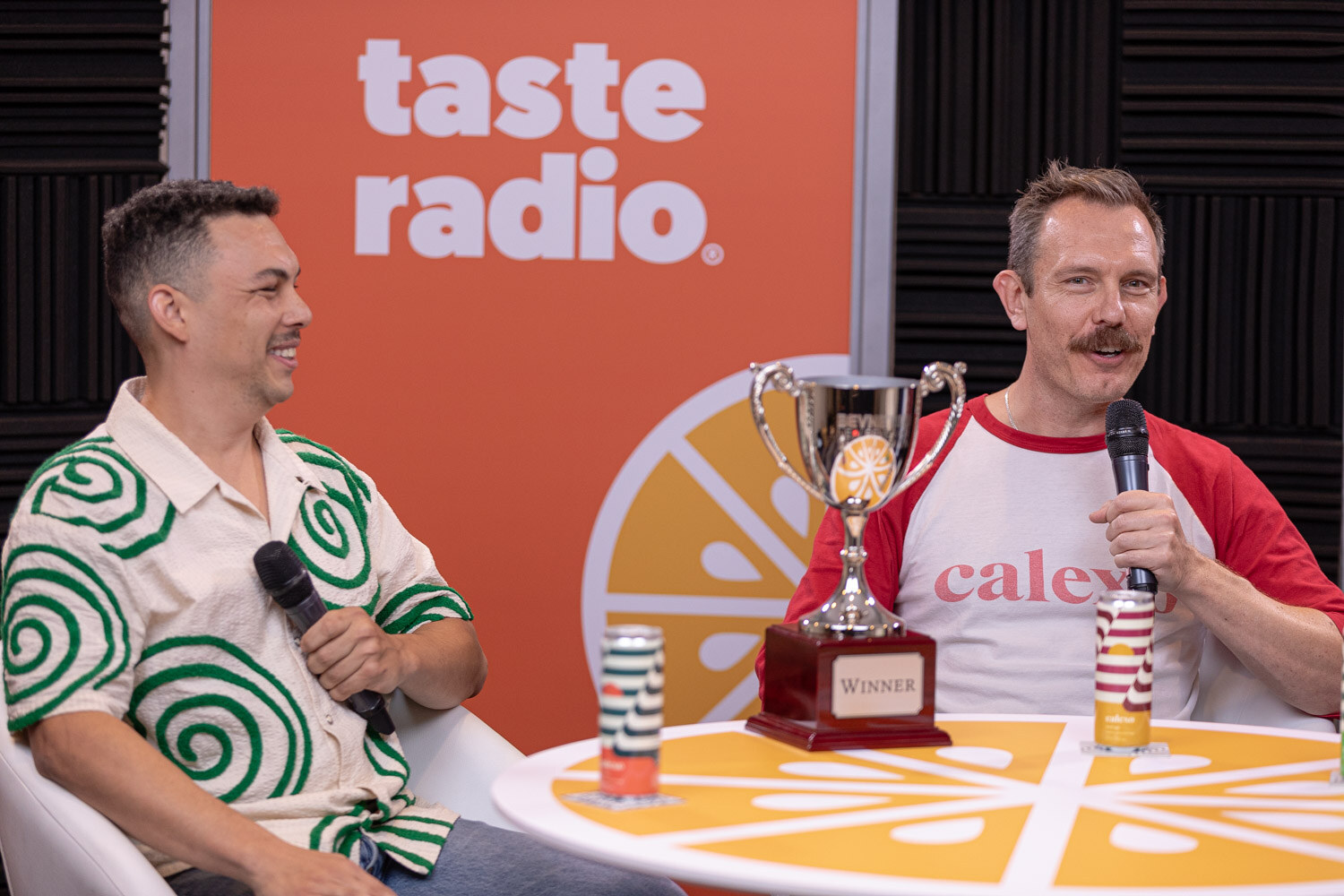 Two men in a Taste Radio studio, one in a green patterned shirt and one in a red and white shirt, with a trophy on the table.