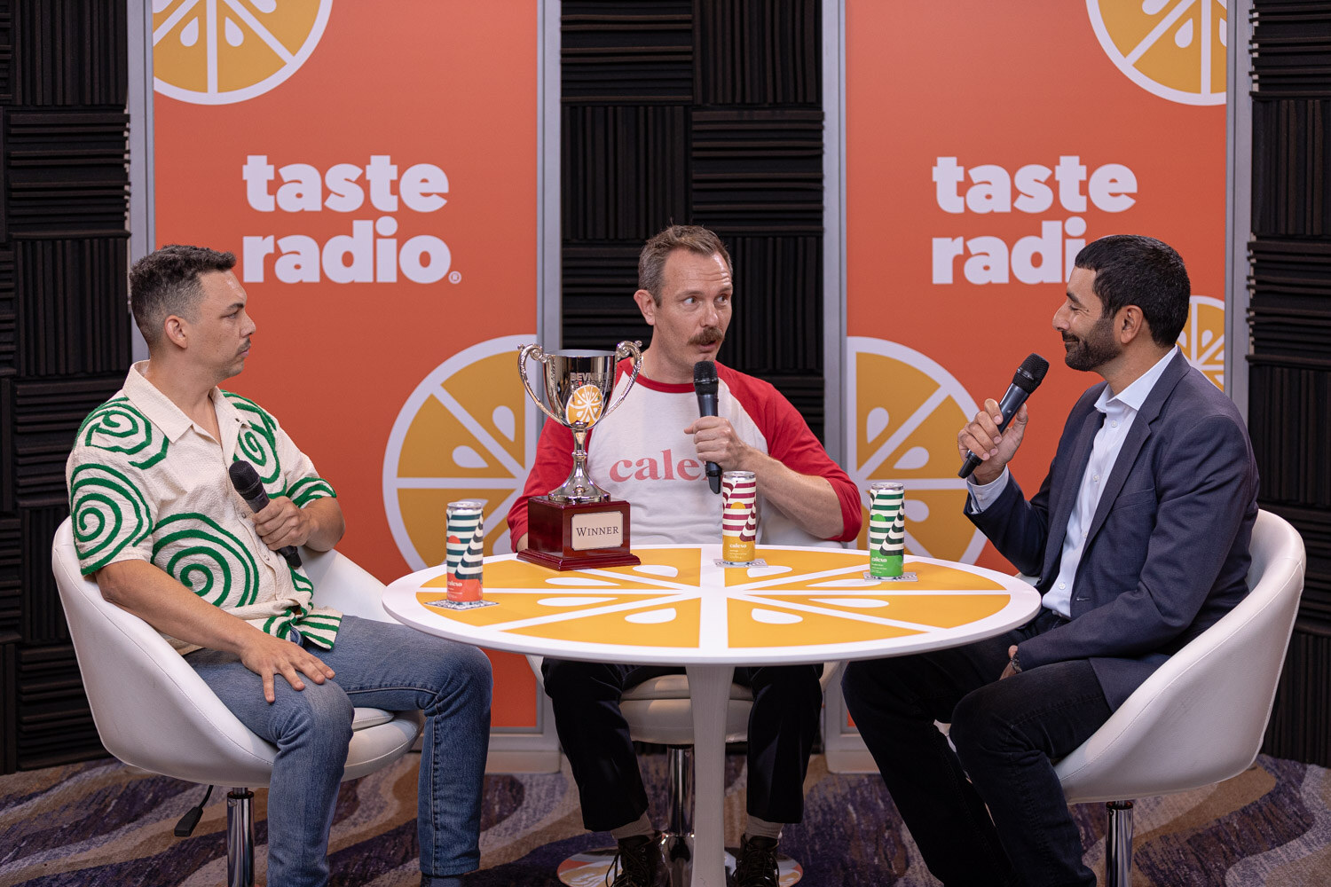 Three men sit around a round studio table with a trophy and four cans in front of a Taste Radio backdrop.
