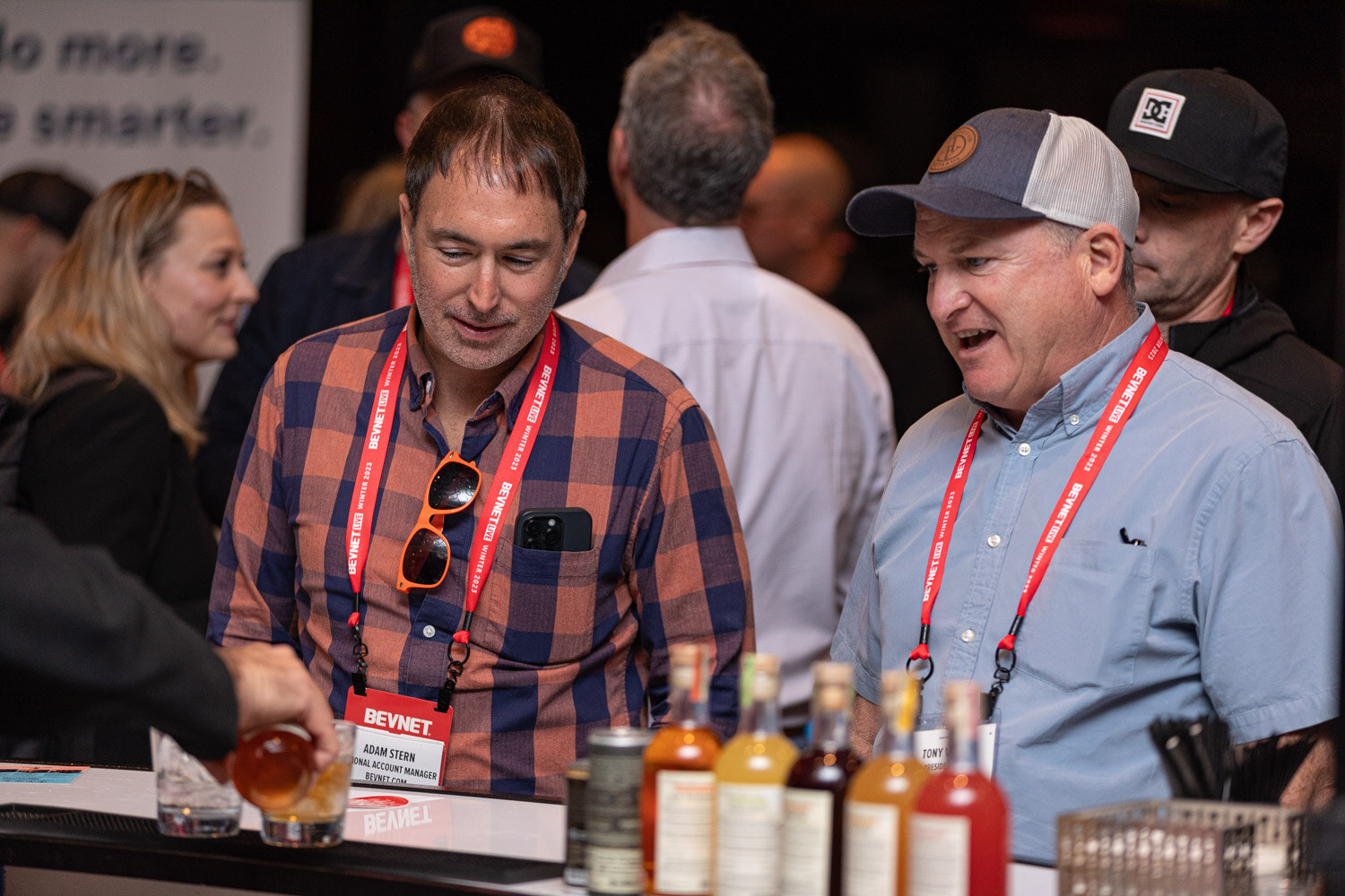 Two men in plaid and blue shirts smile over a bar with bottles of cocktails.