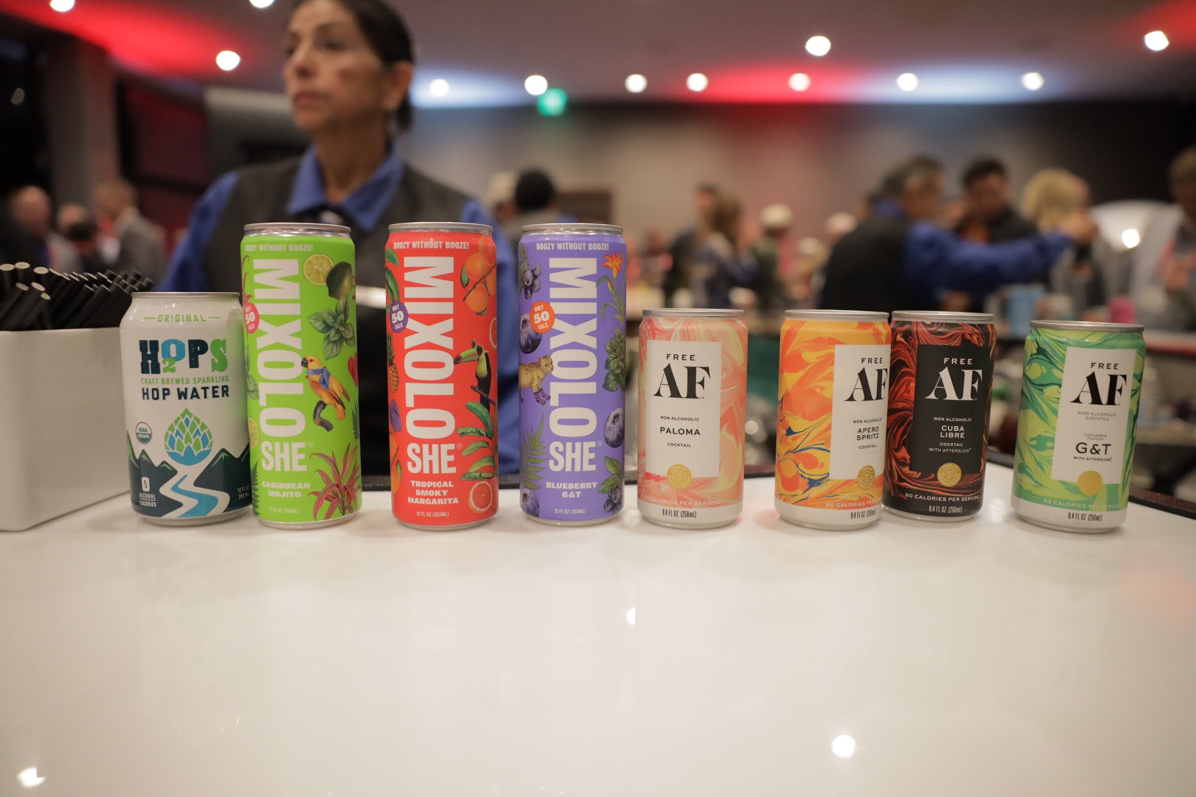 A row of colorful beverage cans on a white table with the party crowd blurred behind it.