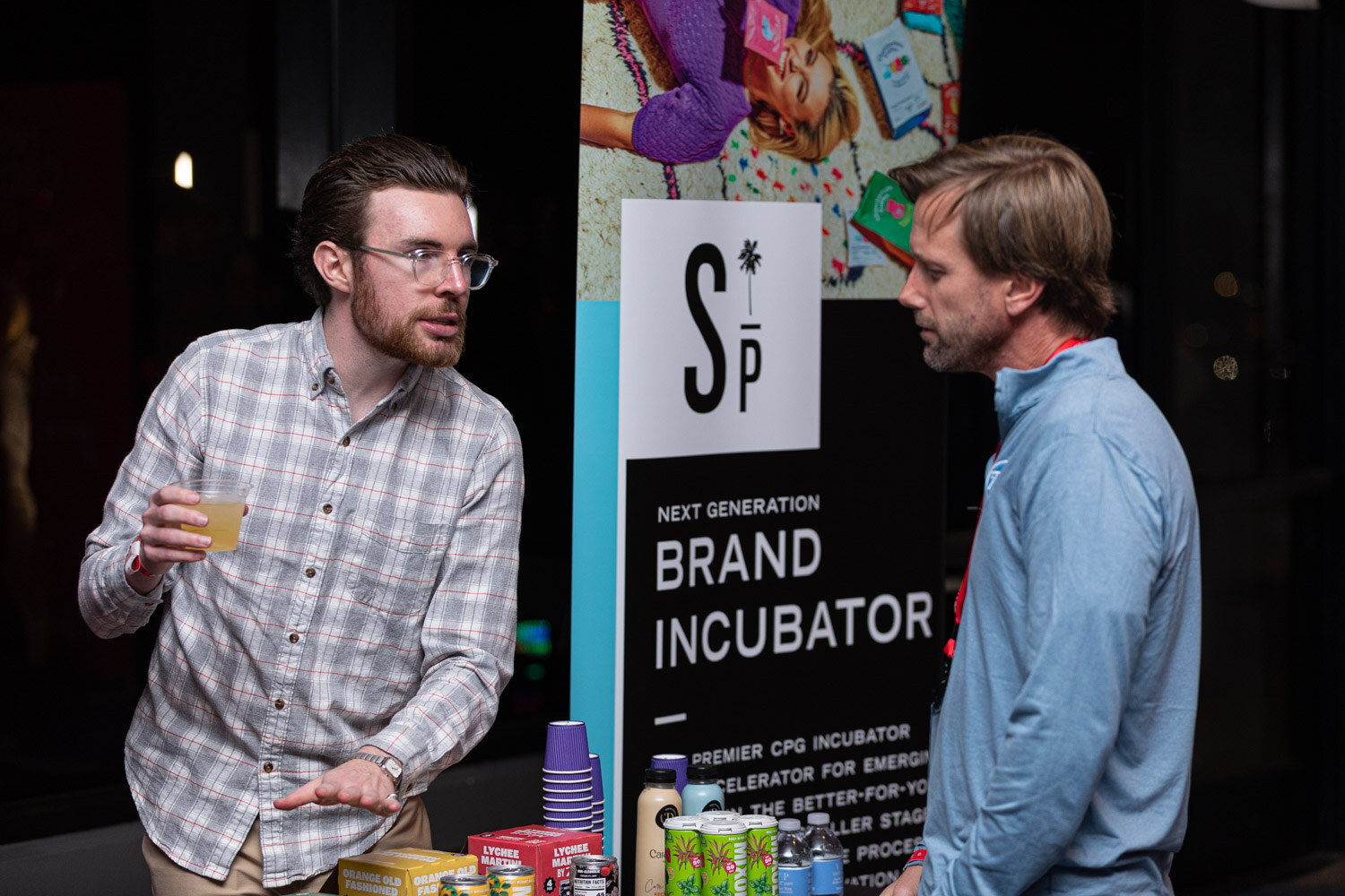 Two men stand in front of a banner reading Brand Incubator holding drinks in plastic cups.