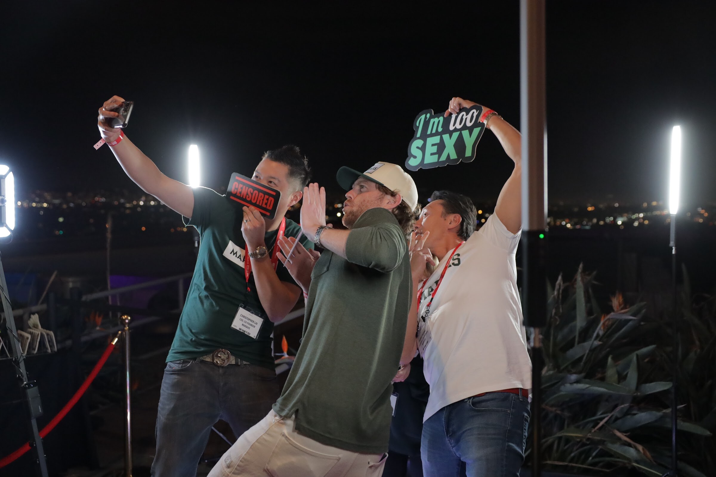 Attendees on a rooftop at night take photos and hold a sign that says I'm too sexy.