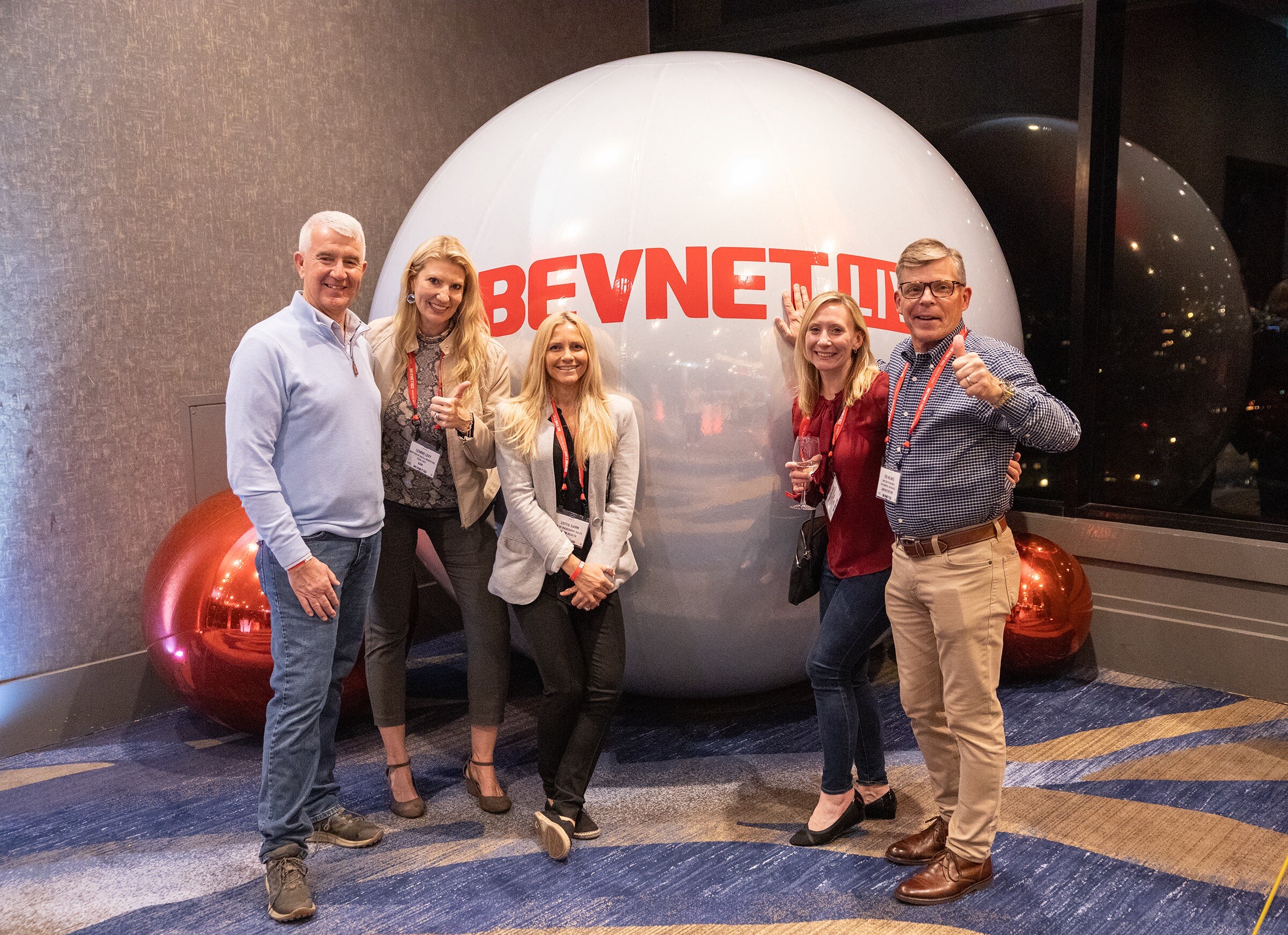 Five attendees in lanyards pose with thumbs up in front of a giant silver ball printed with BevNET Live.